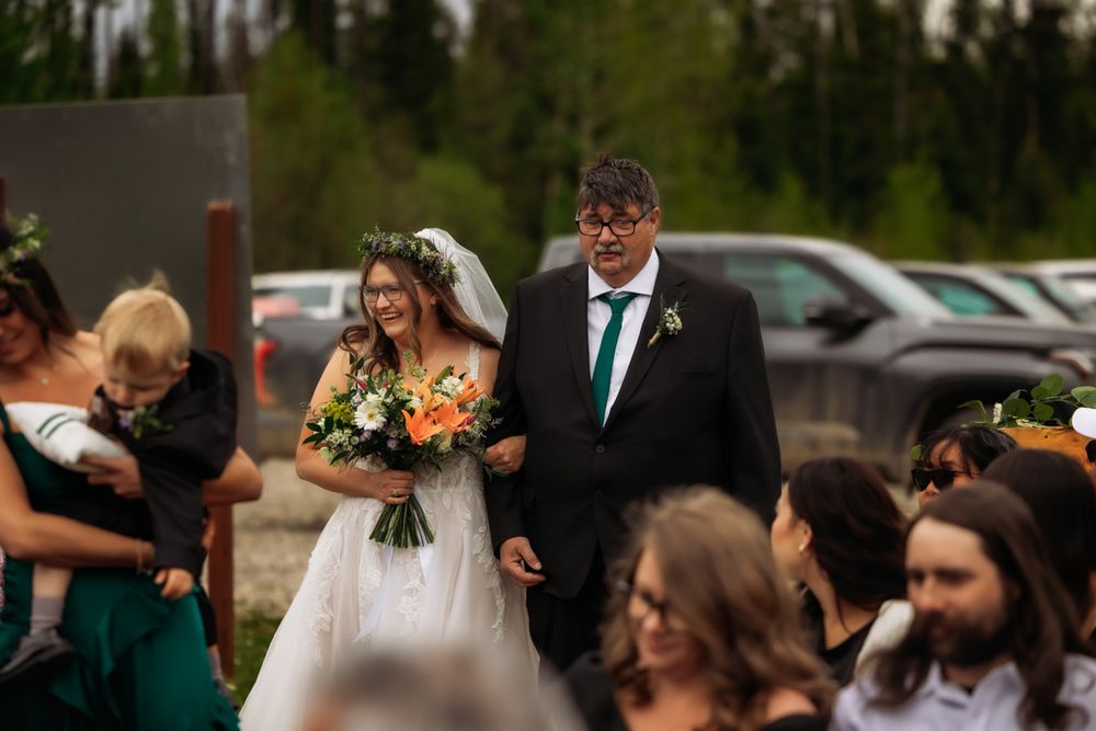 Bride walks down aisle with escort holding colorful bouquet at outdoor wedding ceremony near trees.