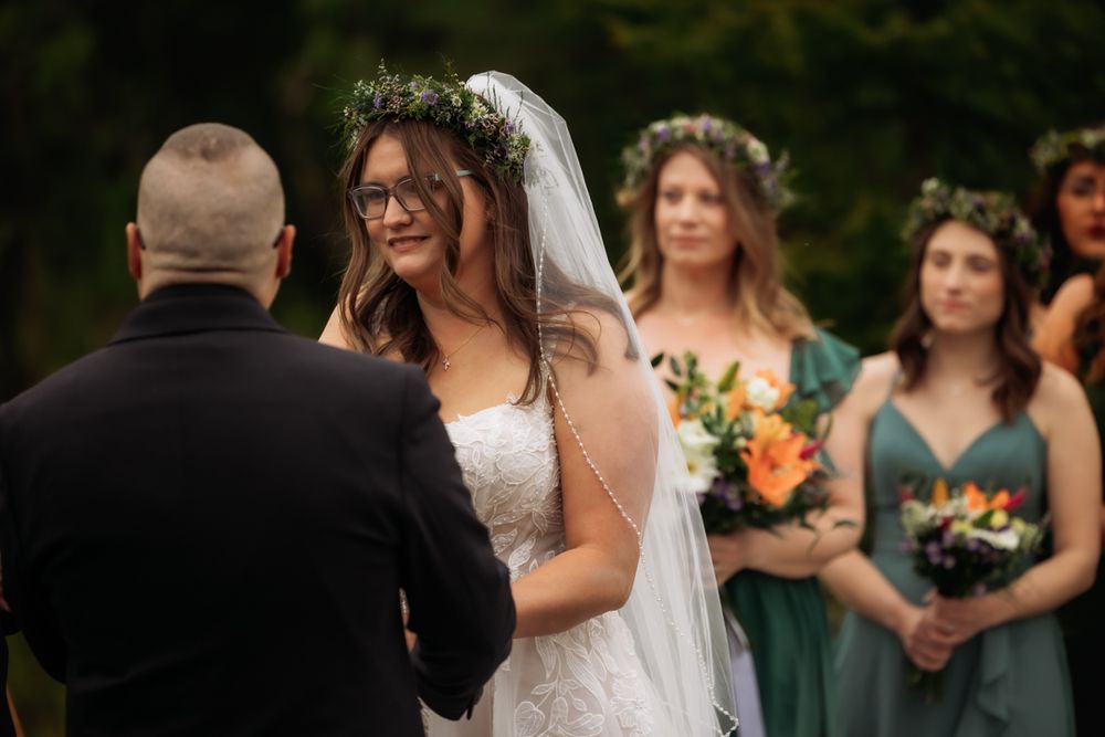 Bride smiling at groom during outdoor wedding ceremony with bridesmaids behind.