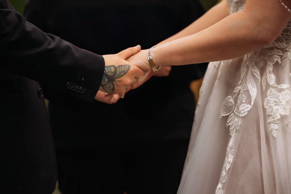 Bride and groom holding hands during wedding ceremony, groom with tattooed arm in black suit, bride in lace gown.