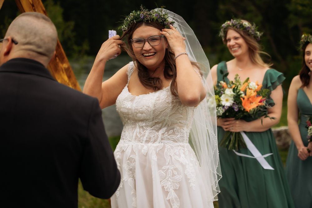 Smiling bride in white lace gown and floral crown adjusts veil at outdoor wedding ceremony with bridesmaids in green.