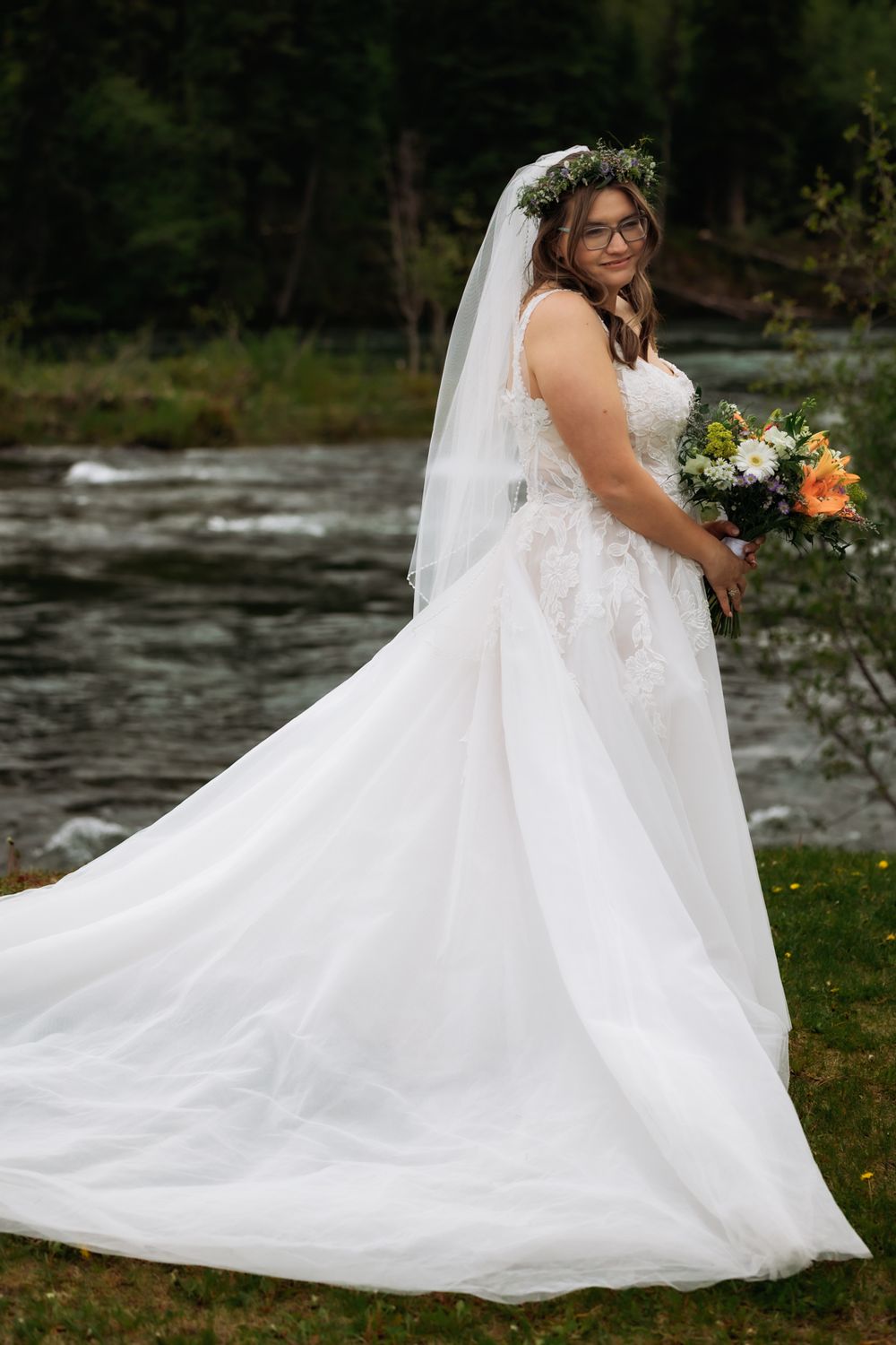 Bride in white lace gown with floral crown holds bouquet beside a flowing river outdoors.