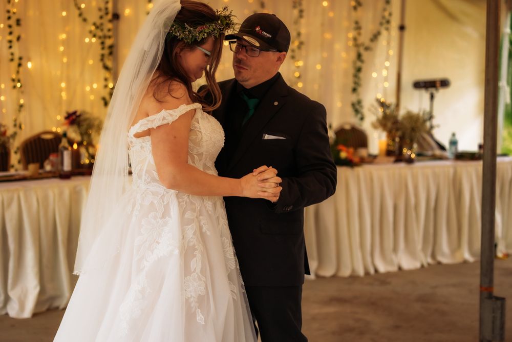 Bride and groom sharing first dance at wedding reception under warm string lights, bride in off-shoulder lace gown.