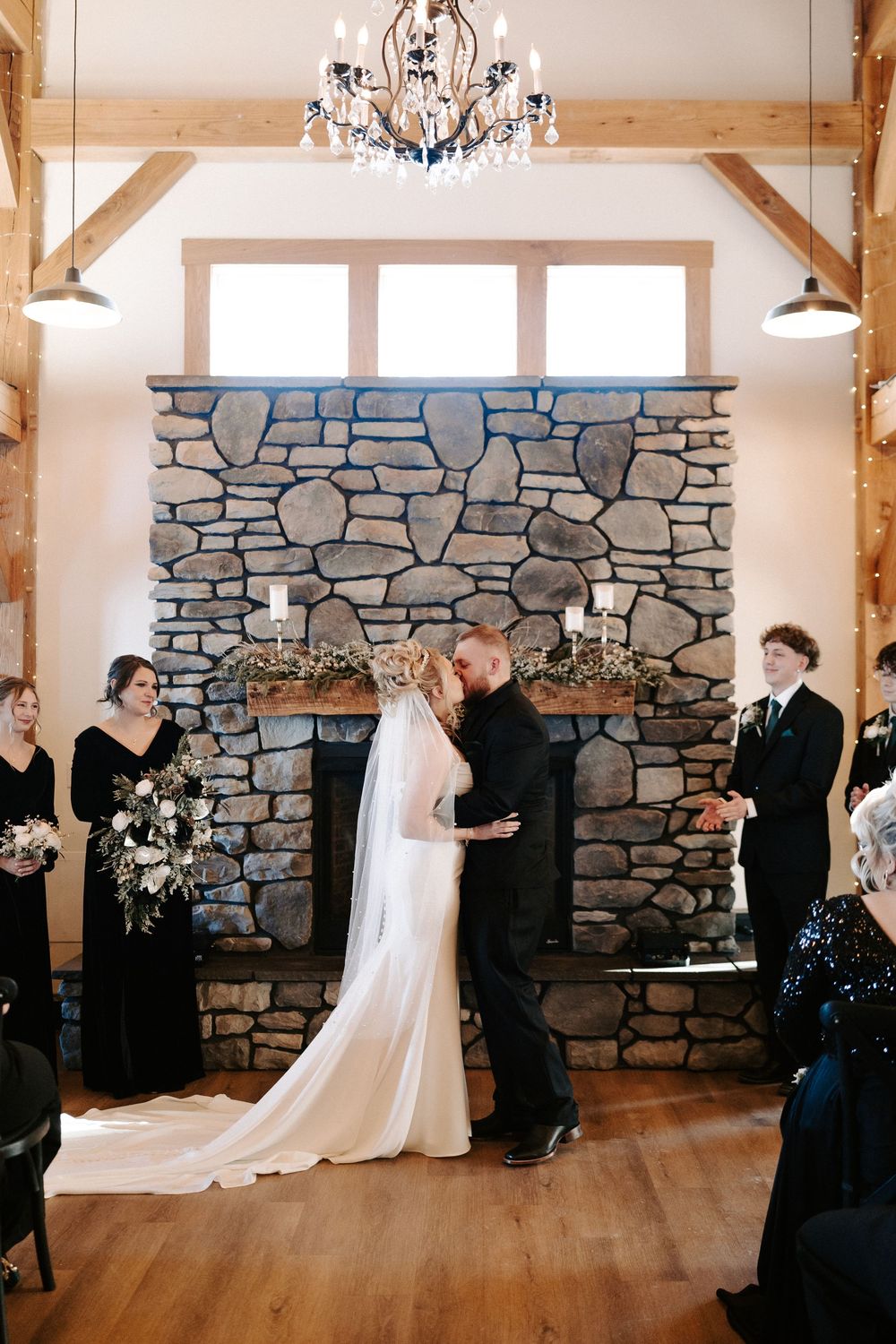Bride and groom sharing first kiss at indoor ceremony before stone fireplace with chandelier.