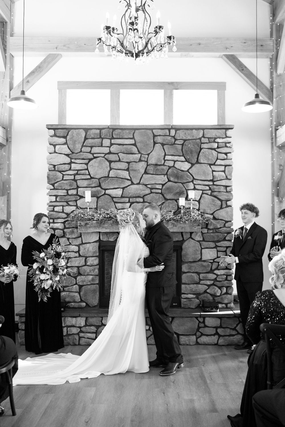 Black and white photo of bride and groom kissing at altar before stone fireplace.