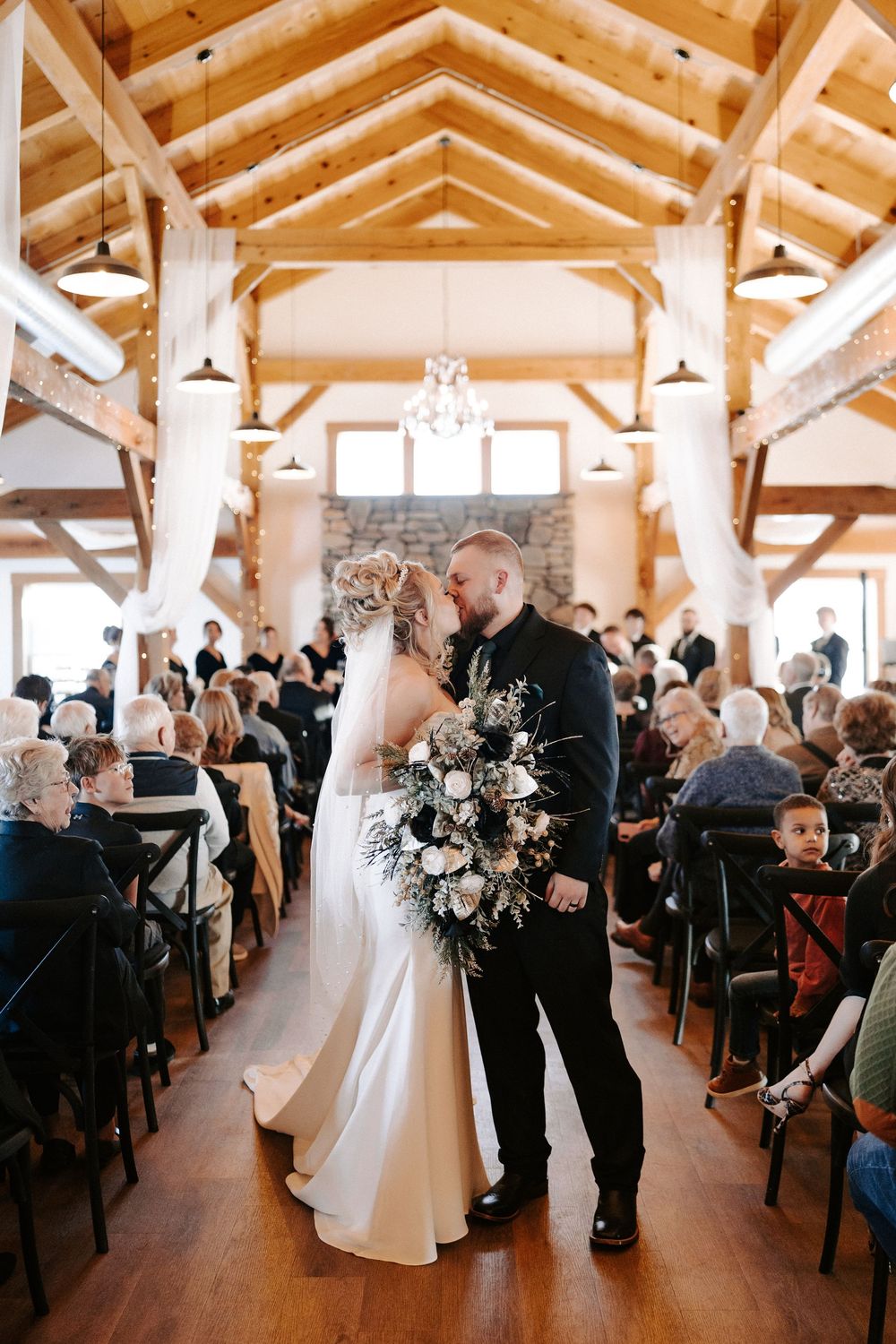 Bride and groom sharing kiss in rustic barn venue aisle with exposed wood beam ceiling.