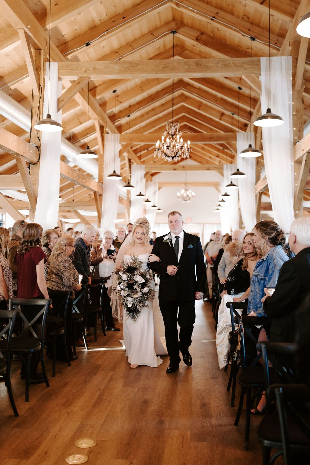 Bride walks down the aisle with escort in elegant rustic barn wedding venue with wood beam ceiling.