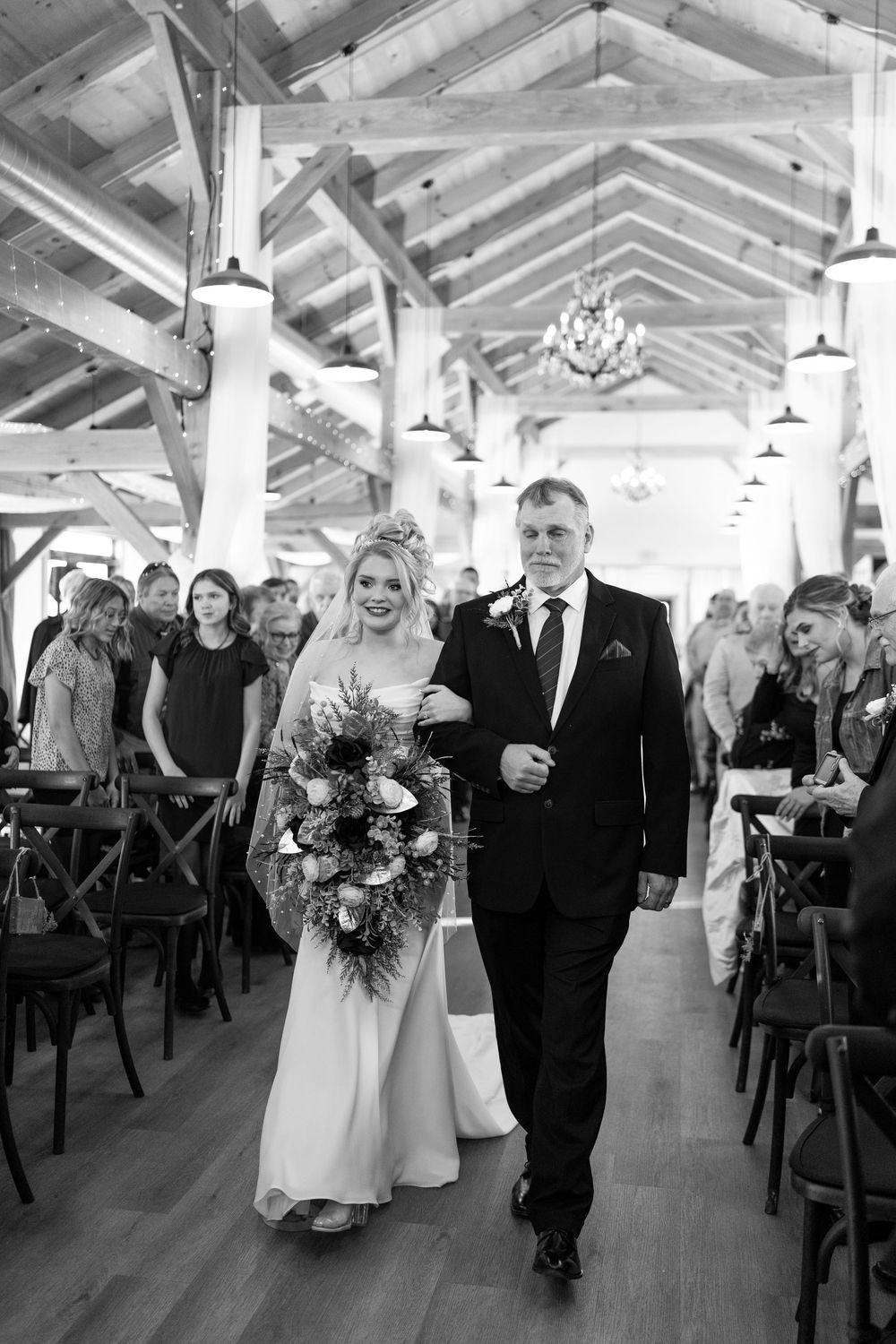 Black and white photo of bride walking down aisle with escort in rustic barn wedding venue.