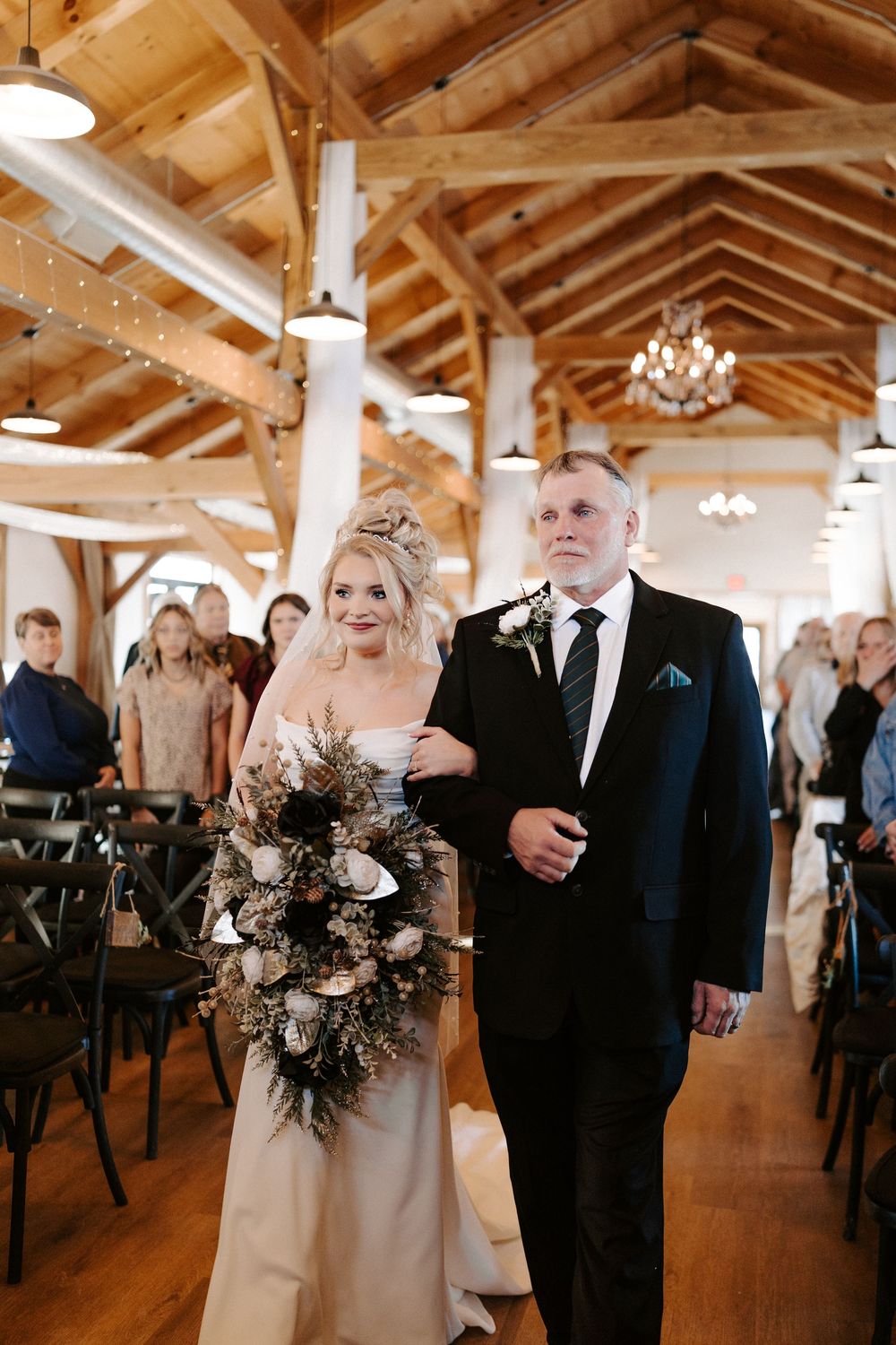 Bride and escort walk down aisle in rustic timber frame wedding venue with exposed wood beams.