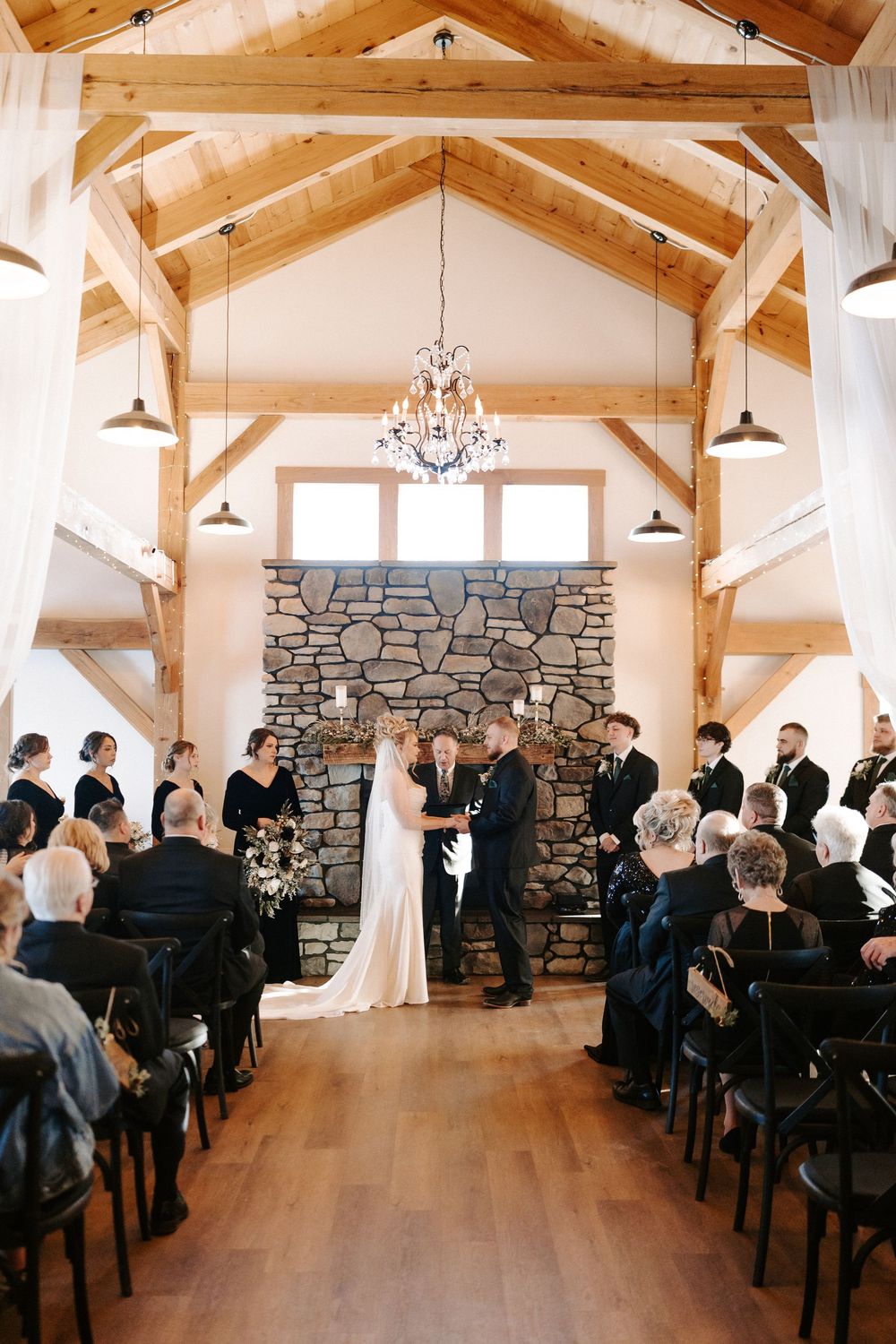 Bride and groom hold hands at altar before stone fireplace in rustic timber frame wedding ceremony.