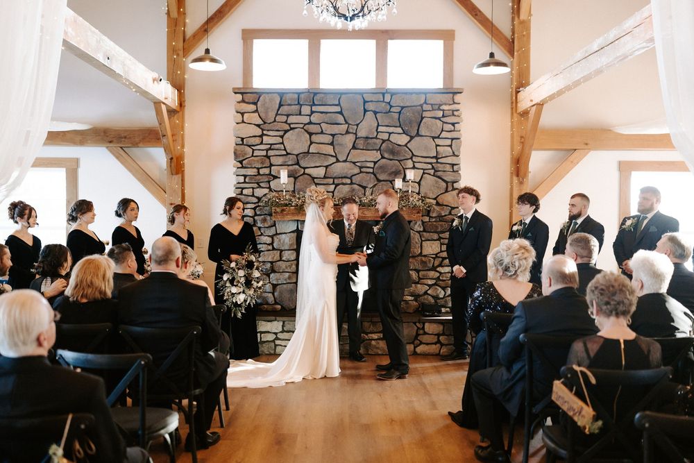 Wedding ceremony at stone fireplace altar with bridal party in black attire in rustic venue.