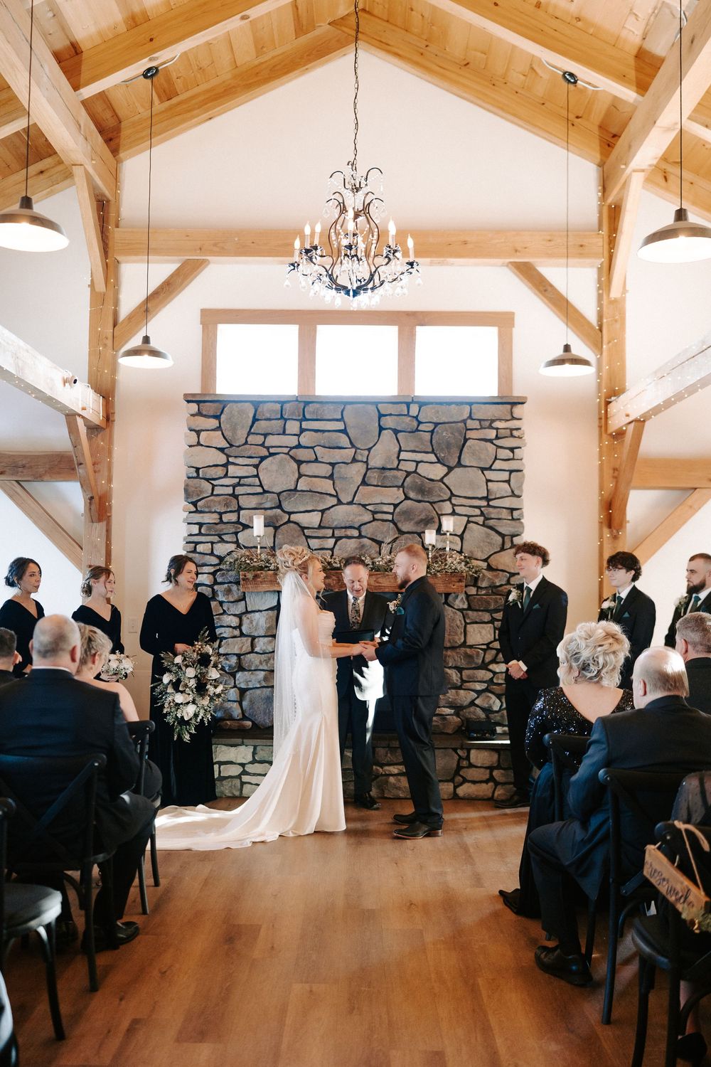 Bride and groom exchange vows before stone fireplace in elegant rustic timber frame wedding venue.