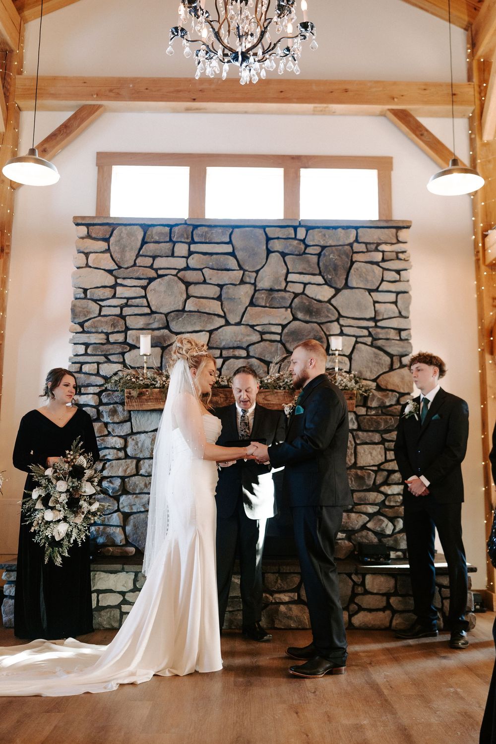 Bride and groom hold hands at stone fireplace altar during rustic barn wedding ceremony.