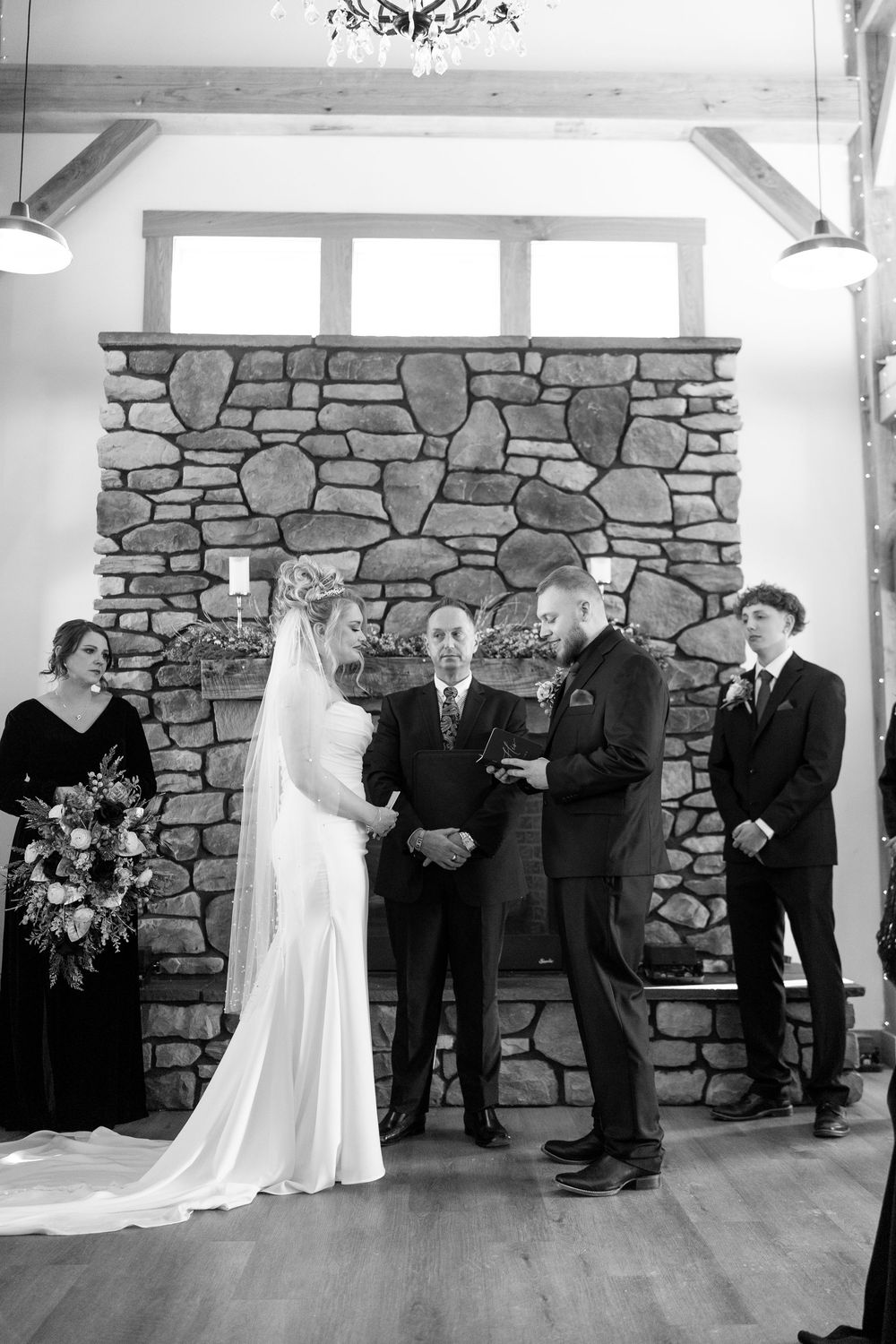 Bride and groom exchange vows at indoor winter wedding ceremony before stone fireplace in black and white photo.