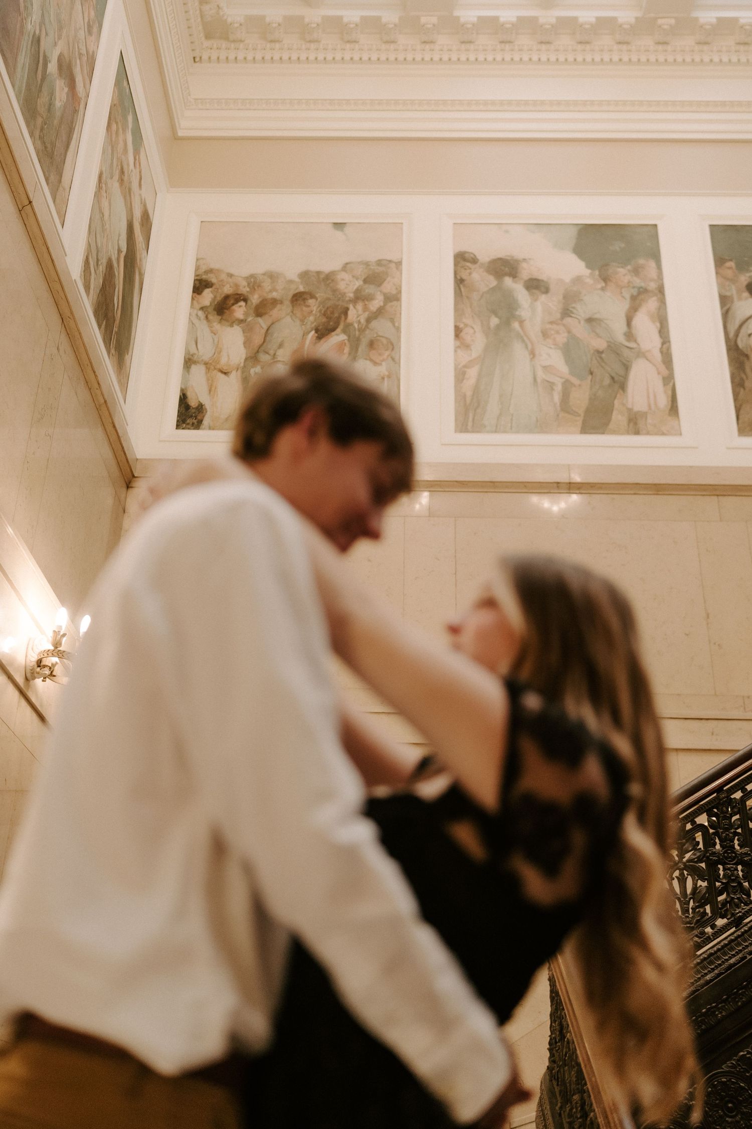 Couple sharing romantic moment on ornate staircase with classical murals and warm lighting behind them.