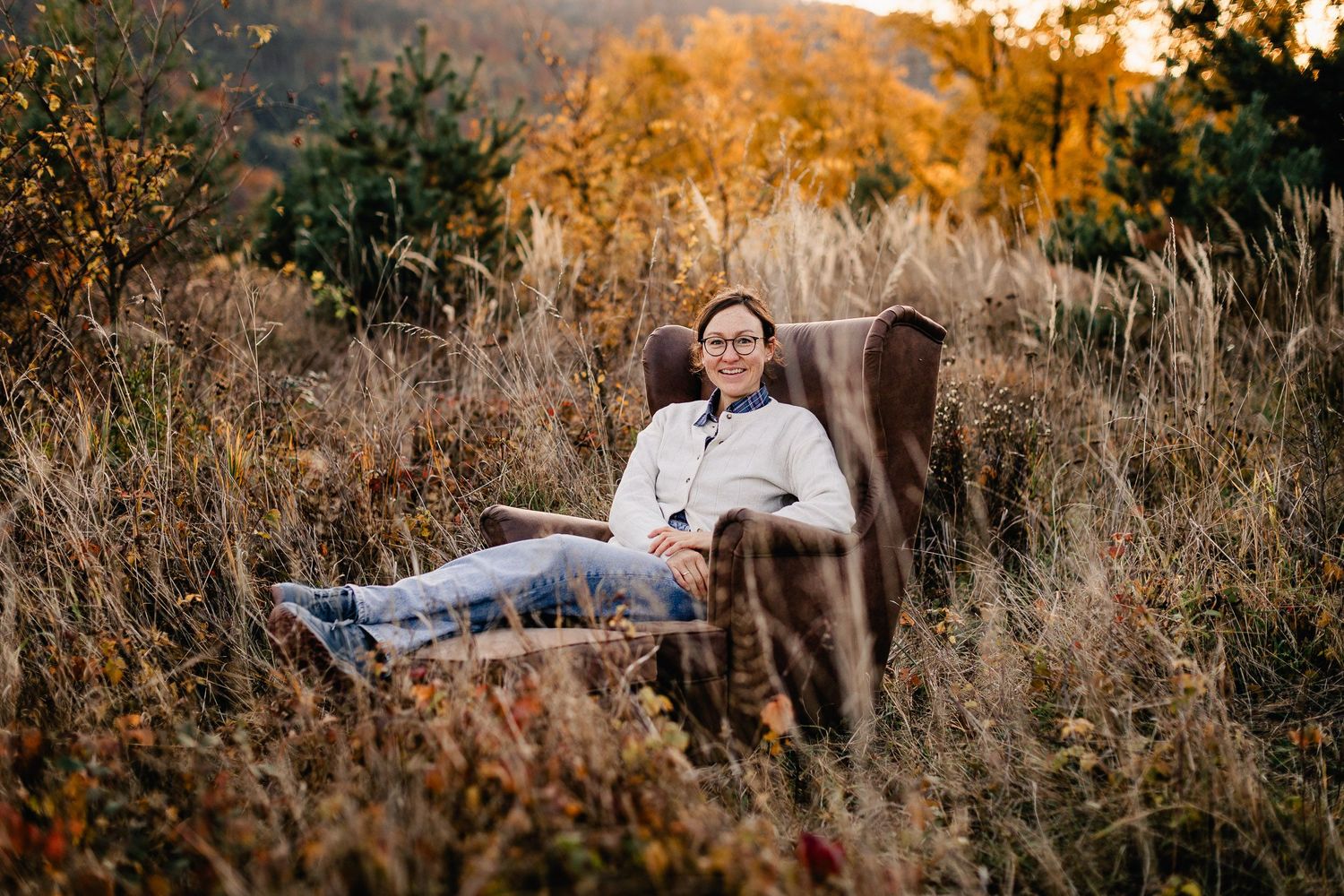 A vintage armchair sits in a golden autumn meadow at sunset with tall grass and wildflowers surrounding it.