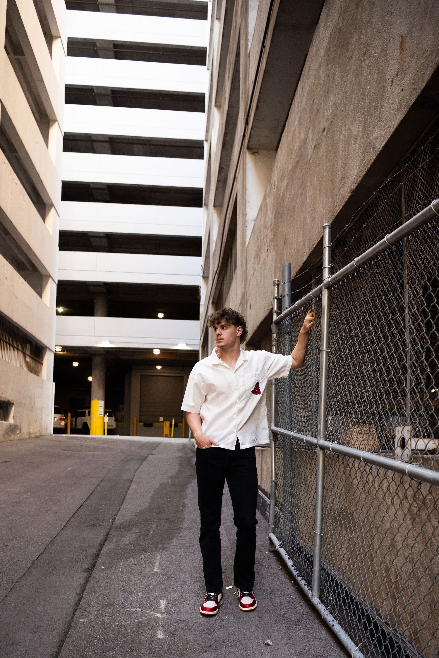 A person in black pants and sneakers walks along a concrete wall with chain link fencing in a parking structure.