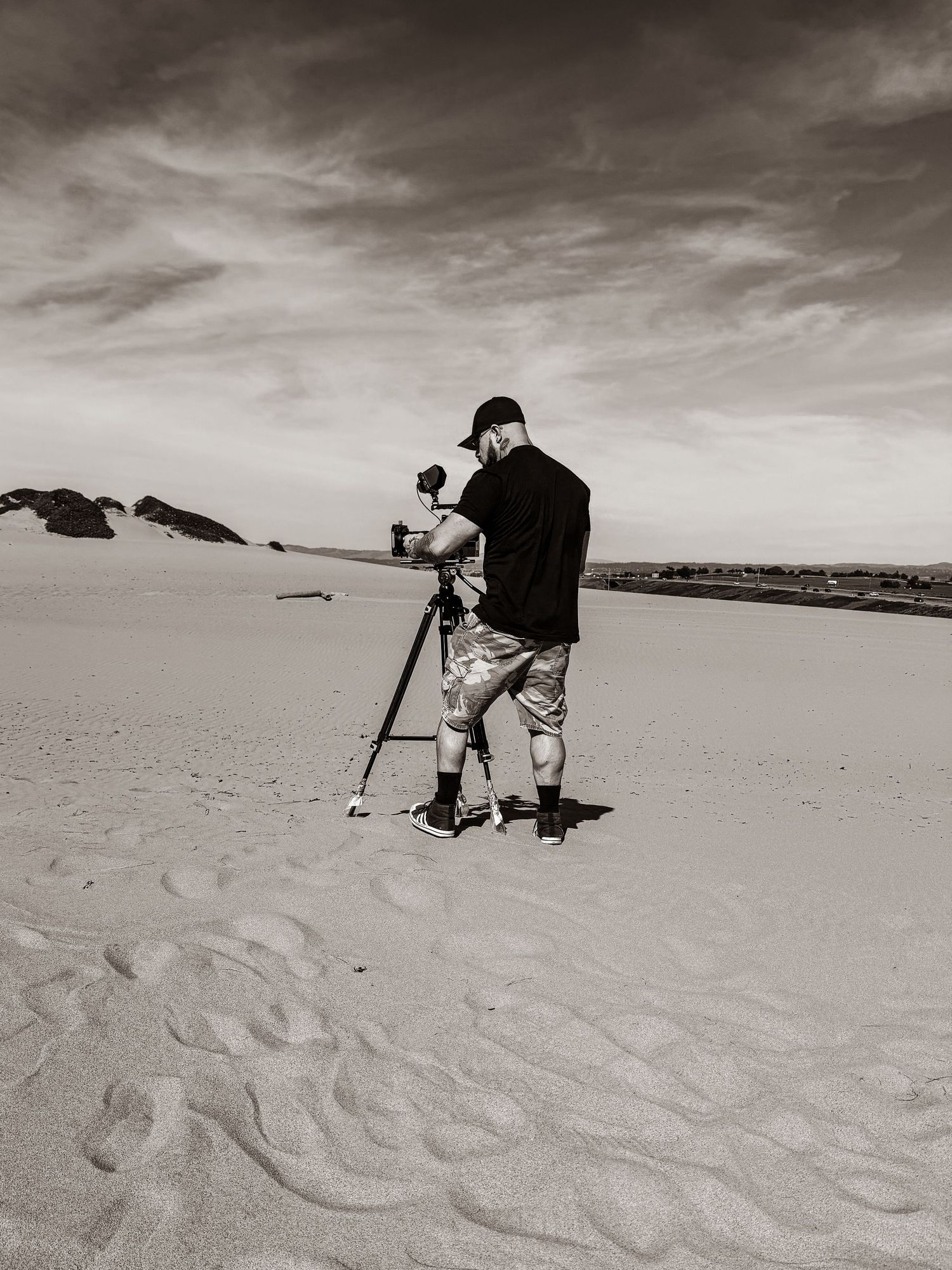 A photographer sets up equipment on a sandy beach under dramatic cloudy skies in black and white.