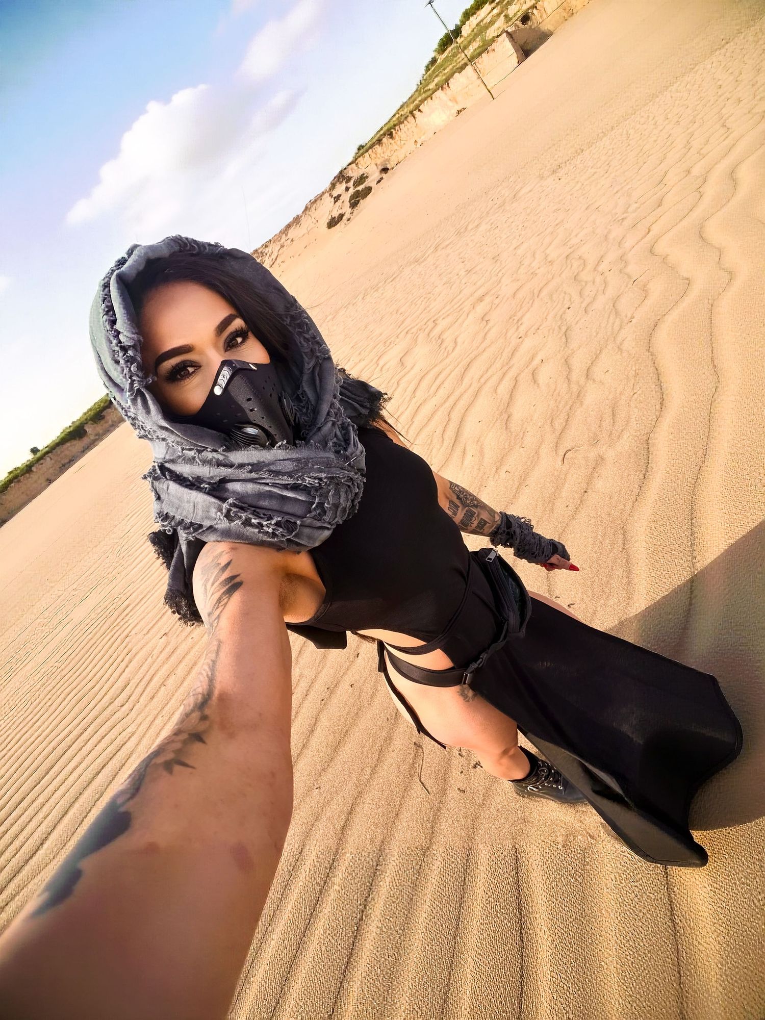 A figure in black attire and head covering poses dramatically against rippling sand dunes in the desert at sunset.