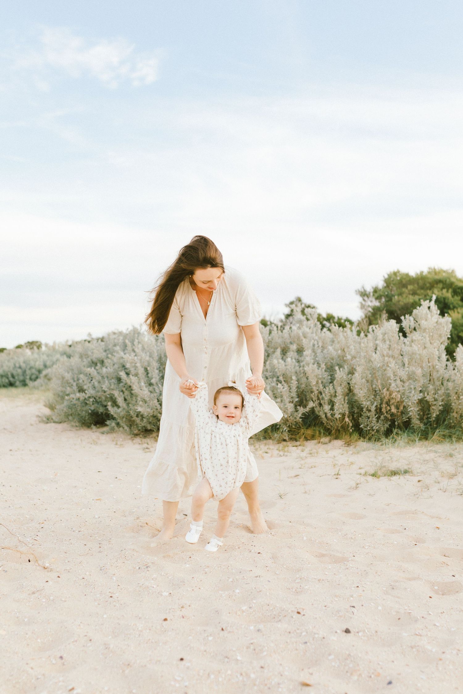 A mother and baby wearing white outfits walk together on a sandy beach path with sage brush in the background.