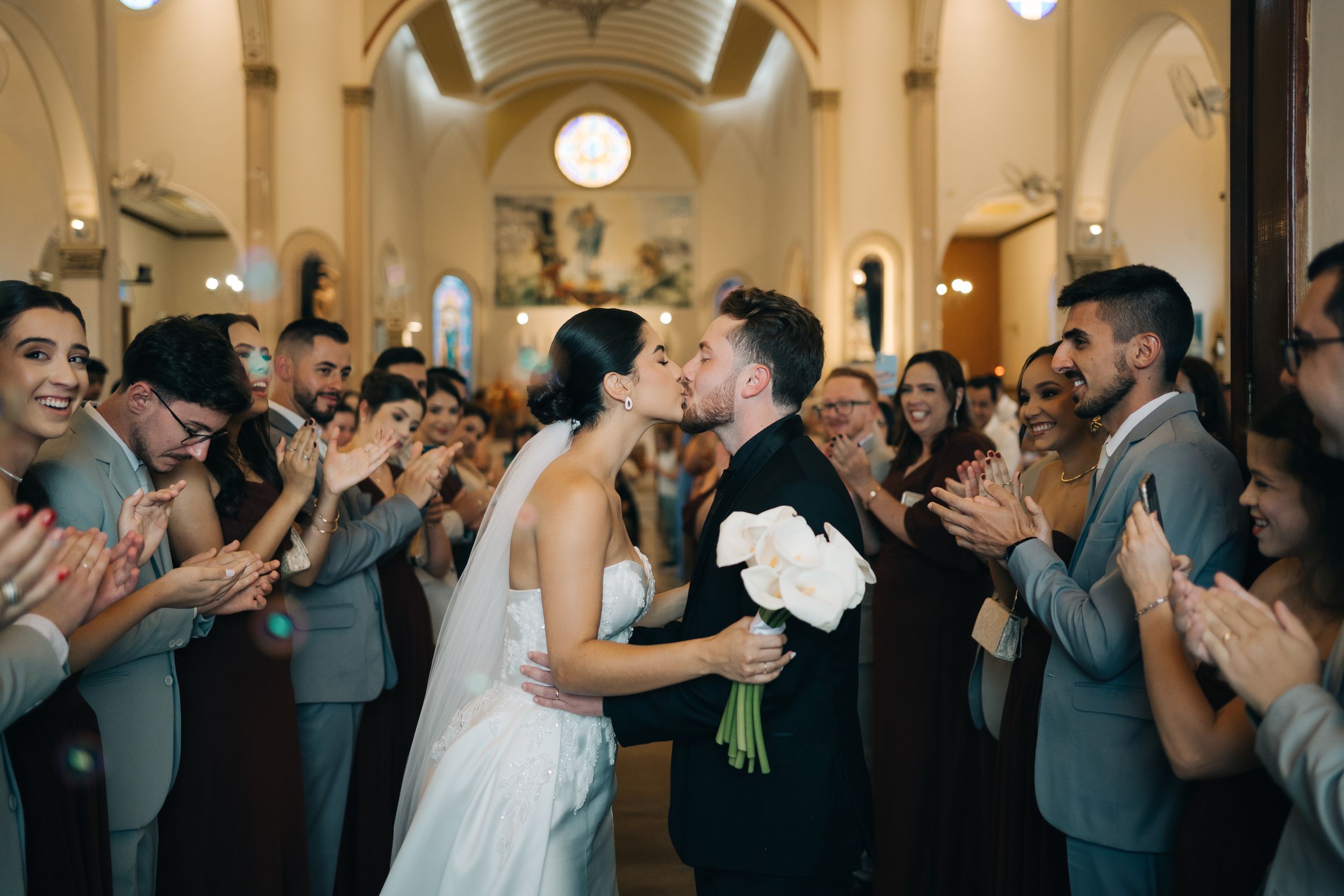 noivos se beijando no corredor da igreja após a cerimonia, com convidados ao fundo aplaudindo e celebrando o casal