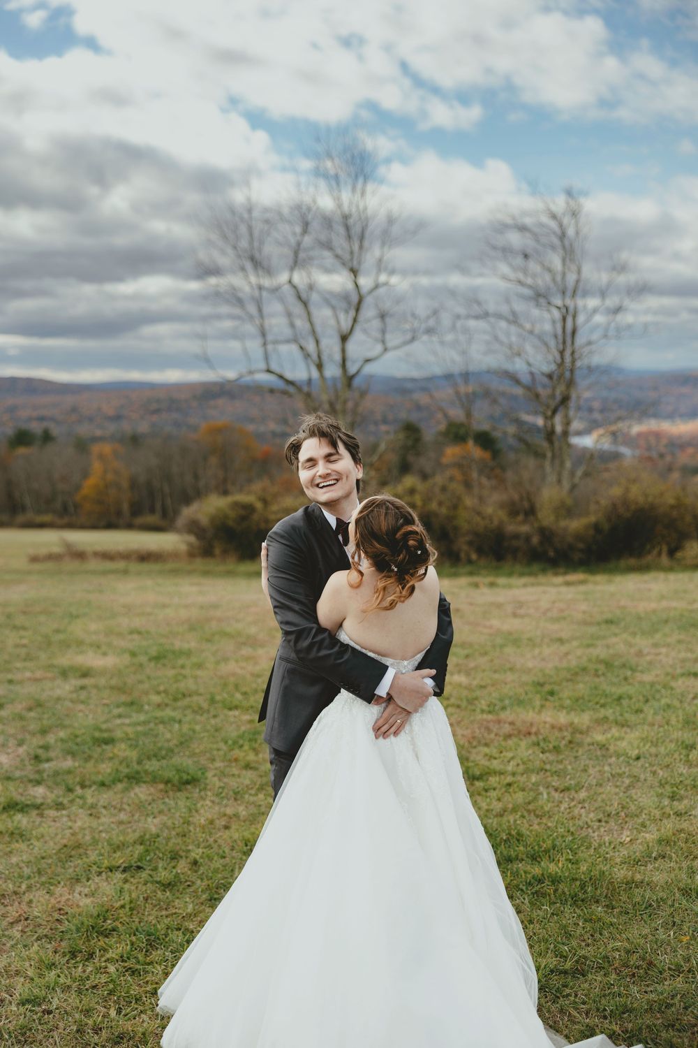 A newlywed couple embraces on a hilltop meadow with autumn mountains and bare trees in the background.