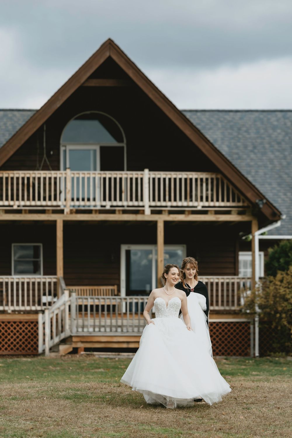 A rustic wooden house with wraparound porches and balconies serves as backdrop for a wedding photoshoot on a cloudy day.