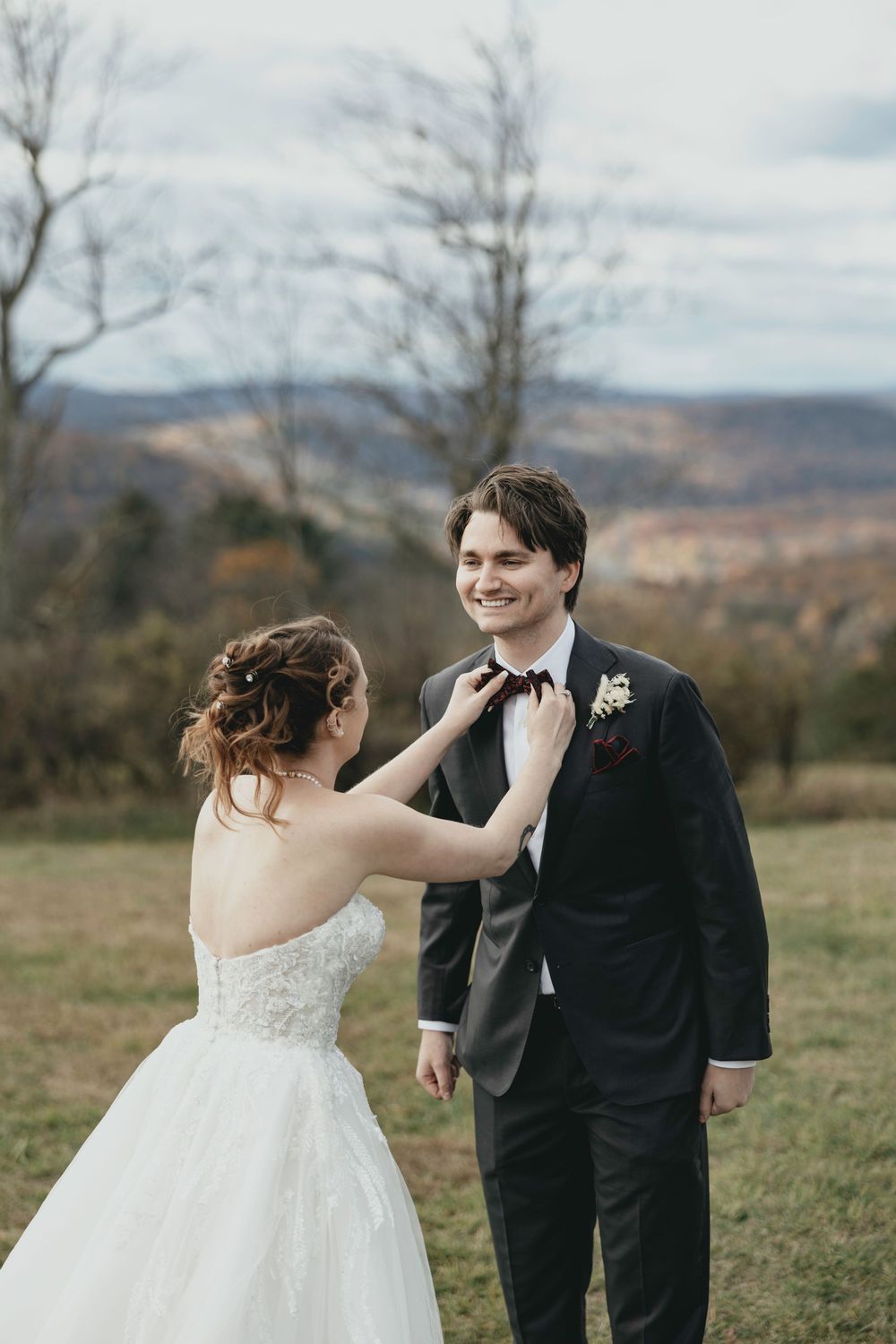 A bride adjusts her groom's bowtie during an intimate outdoor wedding moment with mountain scenery.