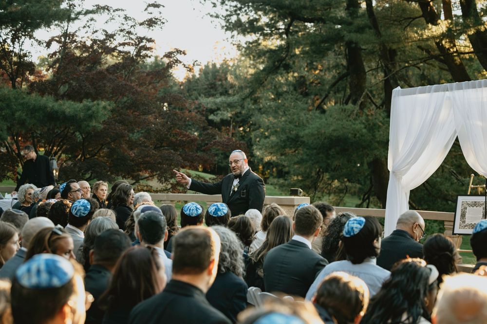 Outdoor wedding ceremony with guests seated in rows facing an altar beneath tall trees at sunset.