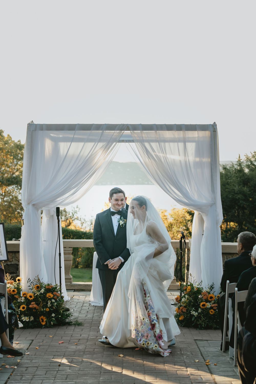 A wedding ceremony setup outdoors with white draped gazebos and sunflowers decorating the aisle at sunset.