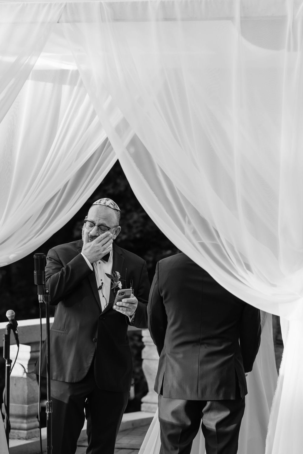 Romantic wedding ceremony with white flowing curtains and guests in formal attire standing in silhouette.