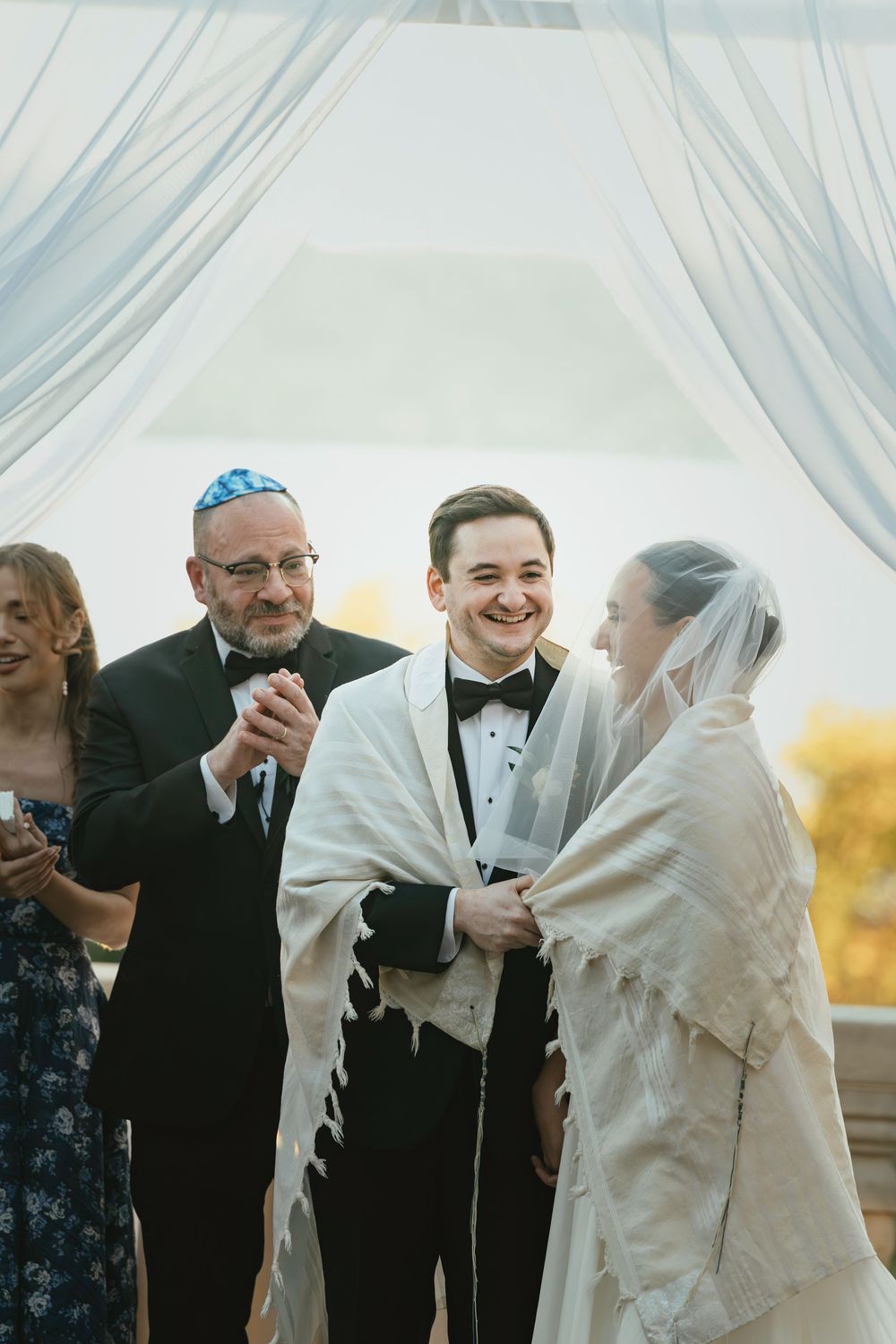 A traditional Jewish wedding ceremony takes place under white fabric draping with autumn colors in the background.