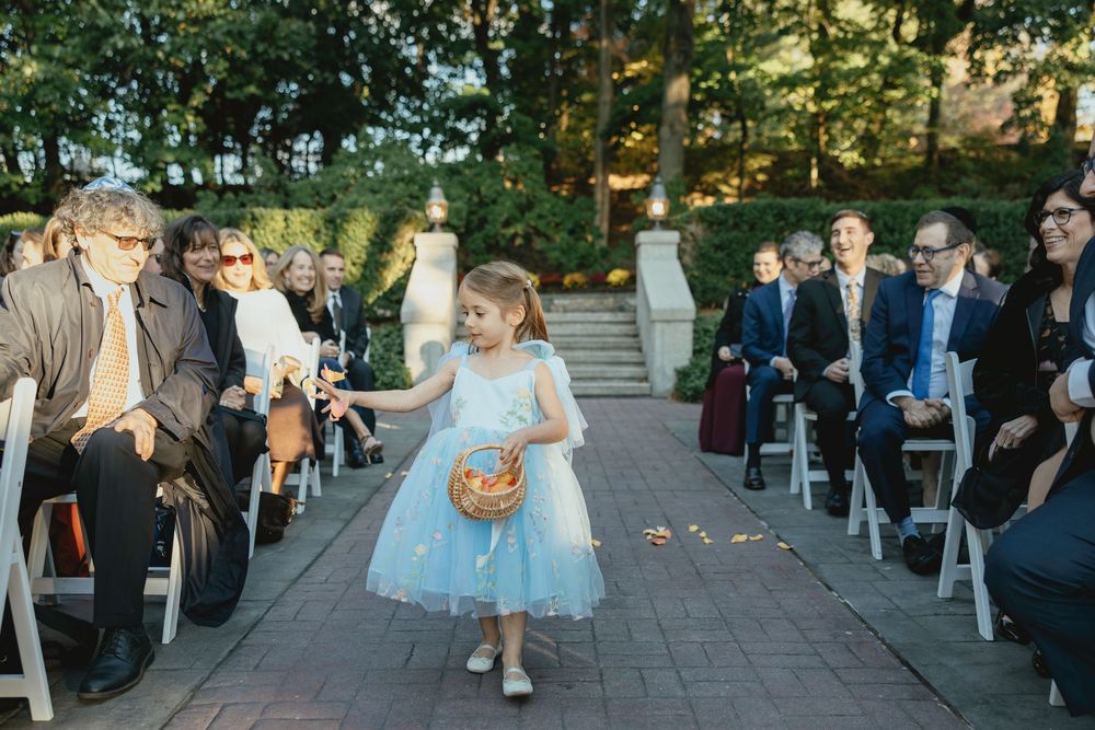 A panoramic view of an outdoor wedding ceremony with guests seated in rows on a stone patio surrounded by trees.