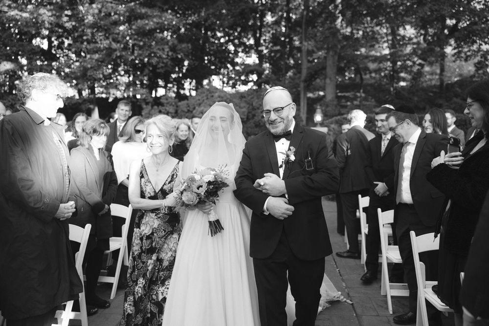 Black and white photo of wedding ceremony guests standing outdoors among trees during daytime celebration.