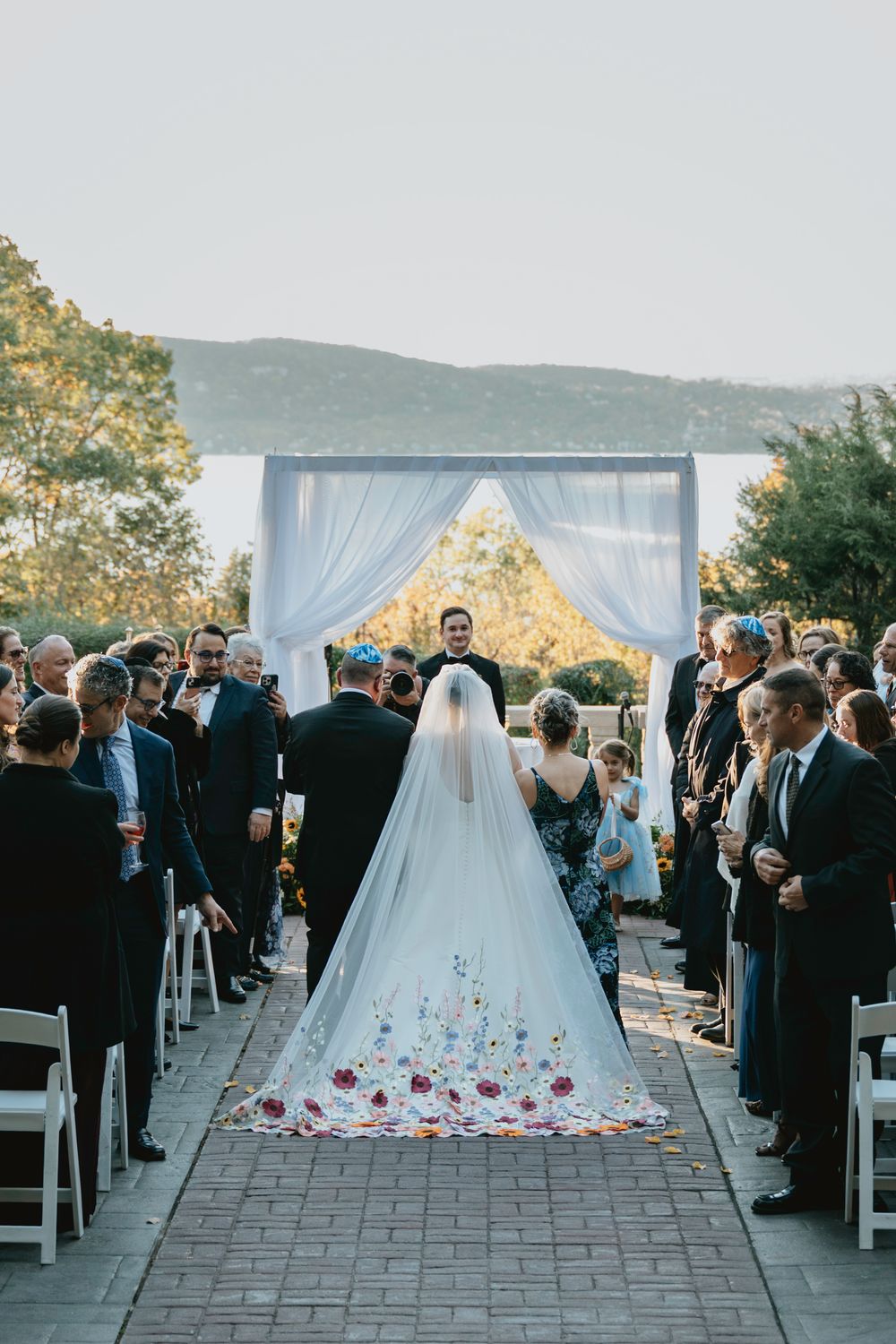 Outdoor wedding ceremony setup with white drapes and rose petals scattered along the aisle at sunset.