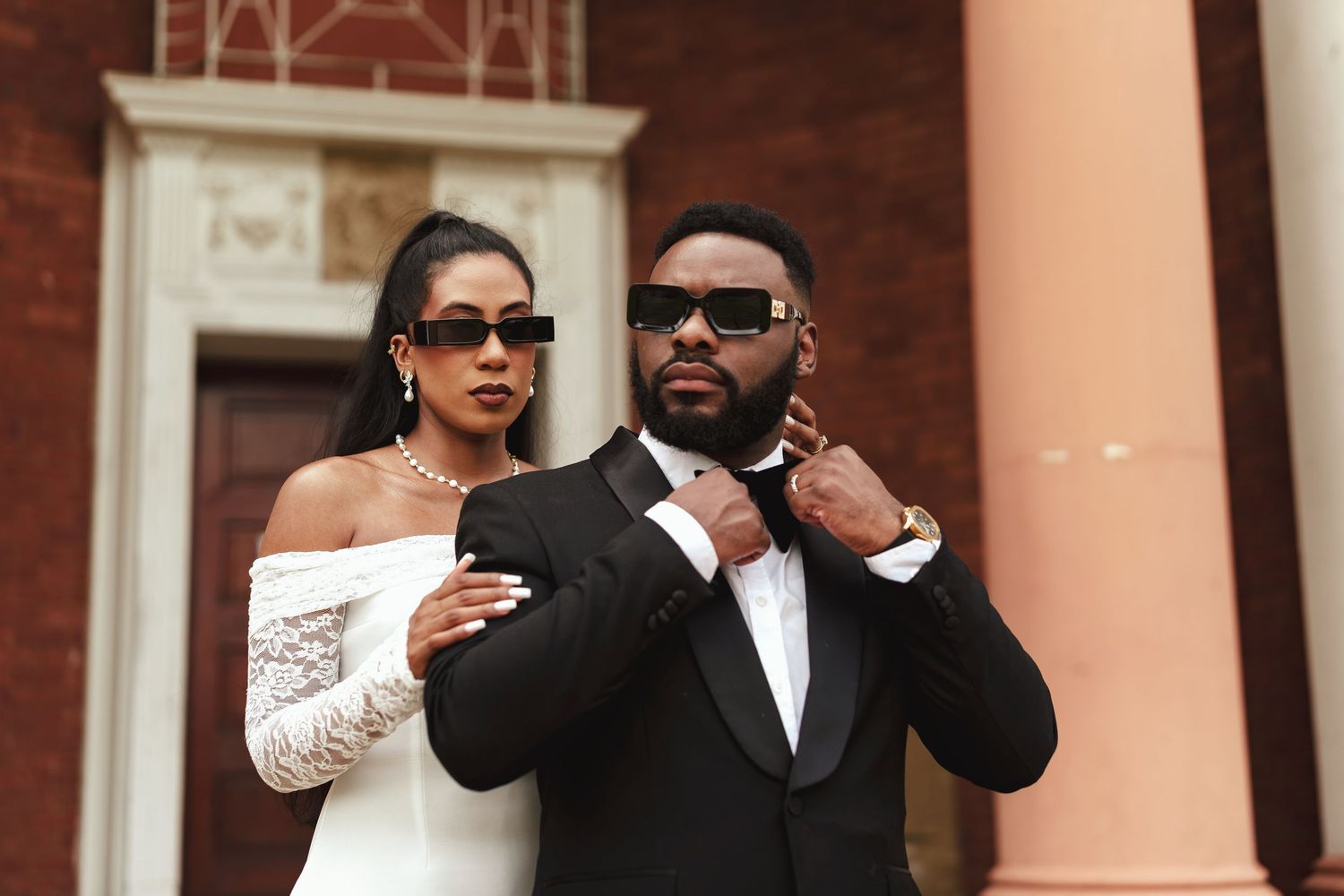 A stylish couple in wedding attire poses outdoors, wearing sunglasses, with the groom in a tuxedo and bride in a white dress.