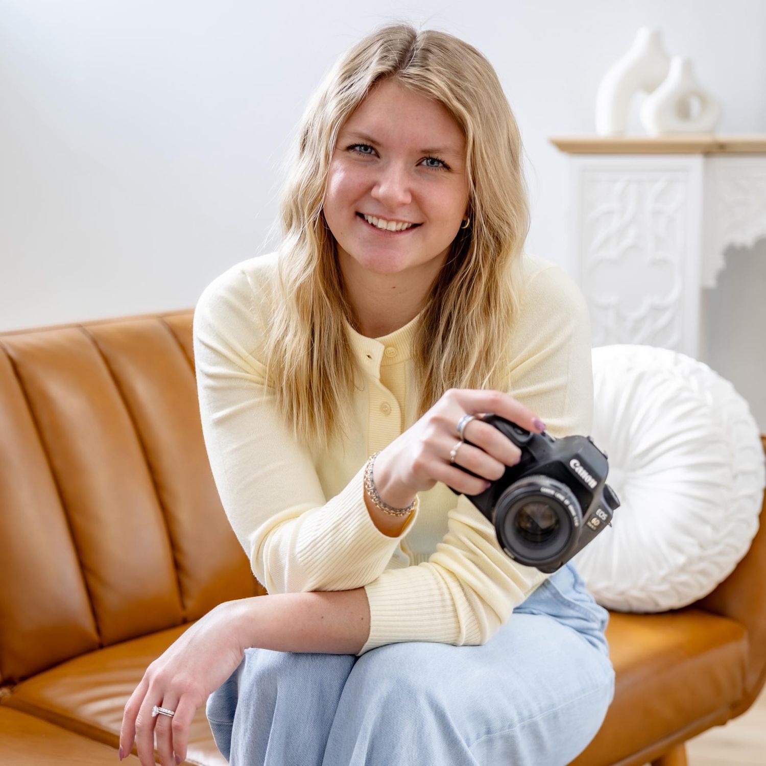 Smiling blonde woman holding a DSLR camera, seated on a brown leather sofa in a bright, modern interior.
