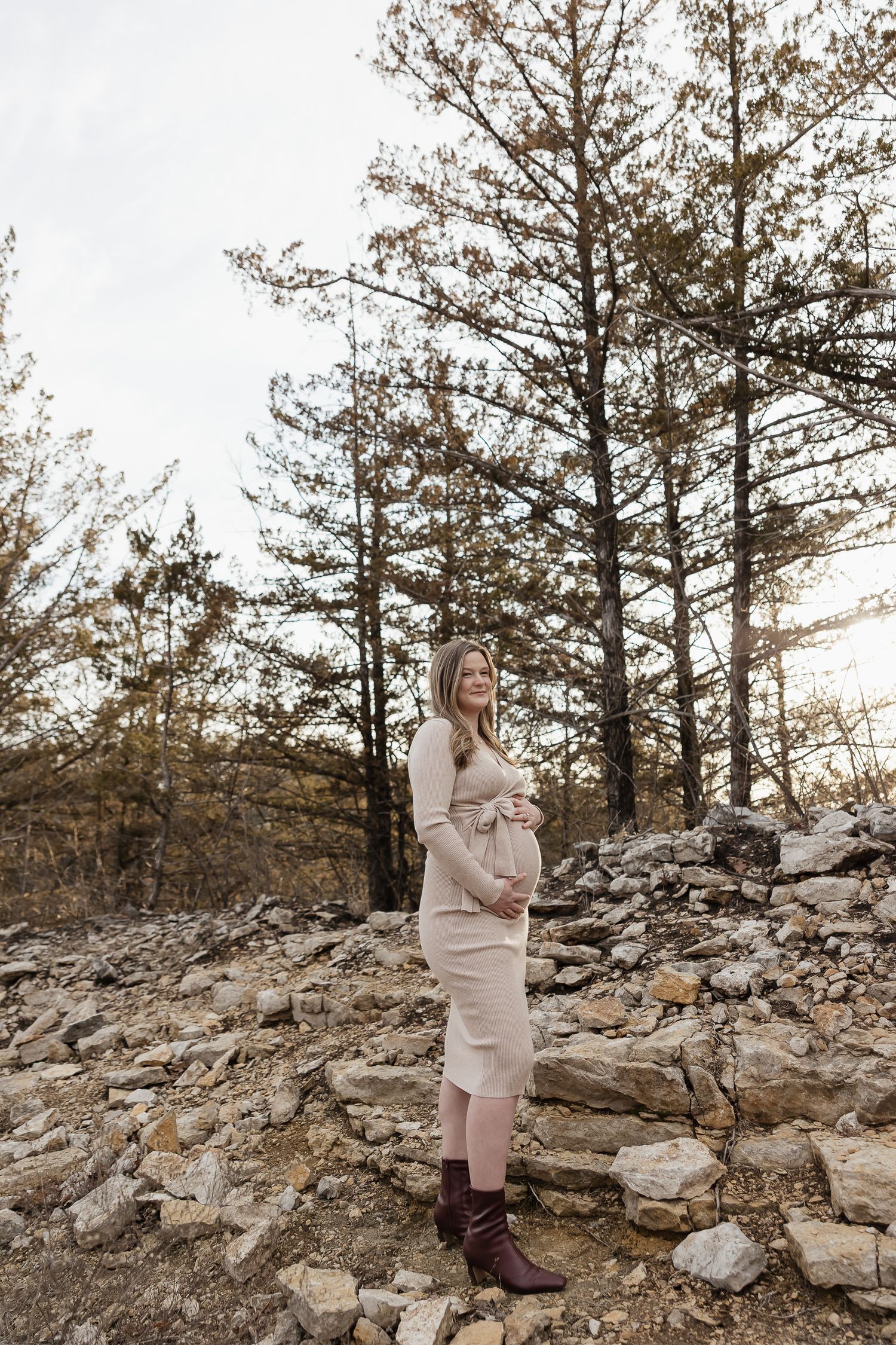 pregnant mother to be holds baby bump in front of pine trees during photo session as date night ideas in kansas city