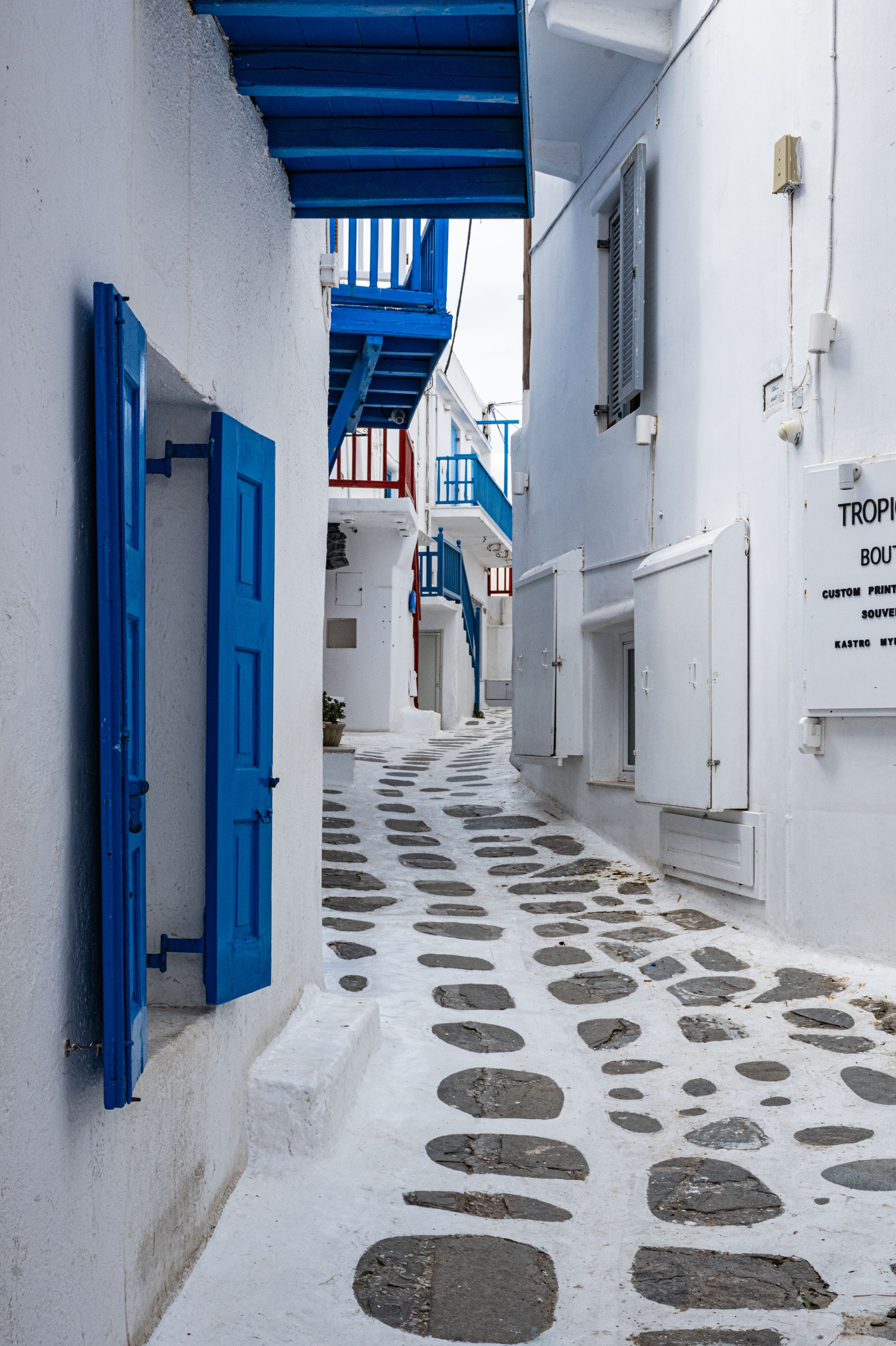 Traditional Greek alley with white walls, blue shutters, and stone paved path in Mykonos.