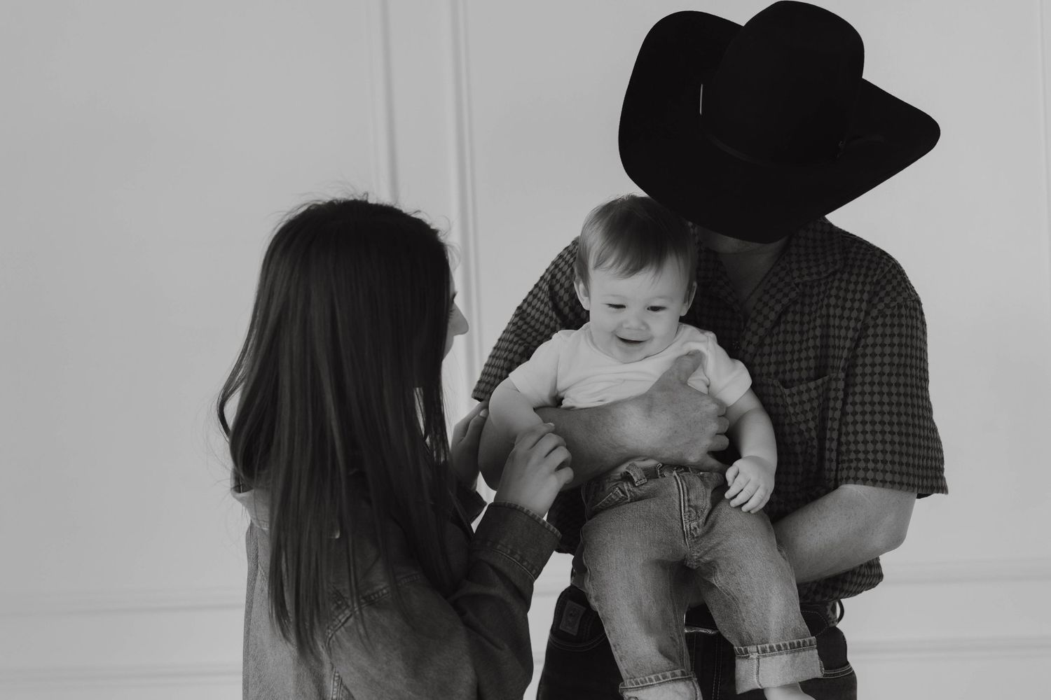 Black and white photo of a man in cowboy hat holding a baby while a woman with long hair reaches out to the child.
