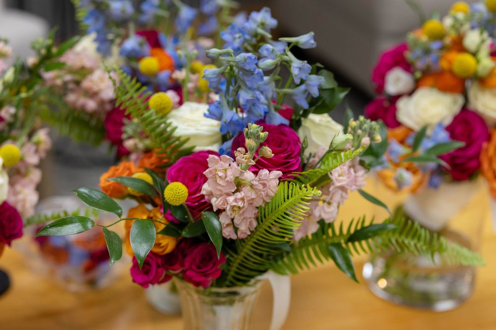 Colorful floral bouquets with pink roses, blue delphiniums, and yellow billy balls arranged in glass vases on a wooden table.