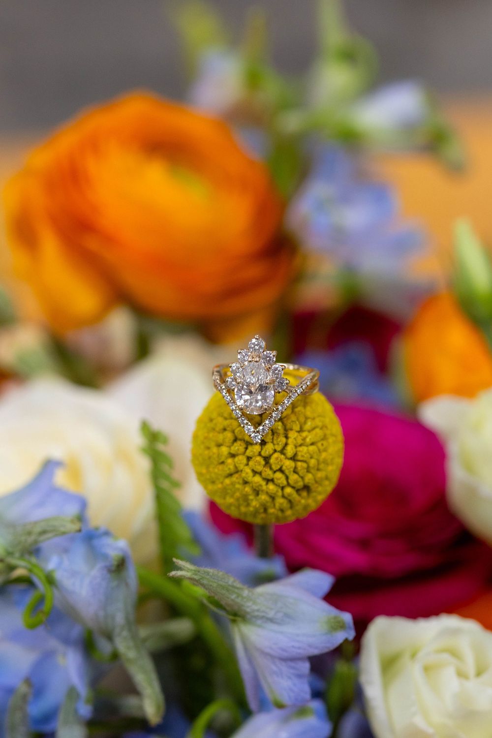Diamond engagement ring balanced on a yellow craspedia flower surrounded by colorful wedding bouquet with orange, pink, and blue blooms.