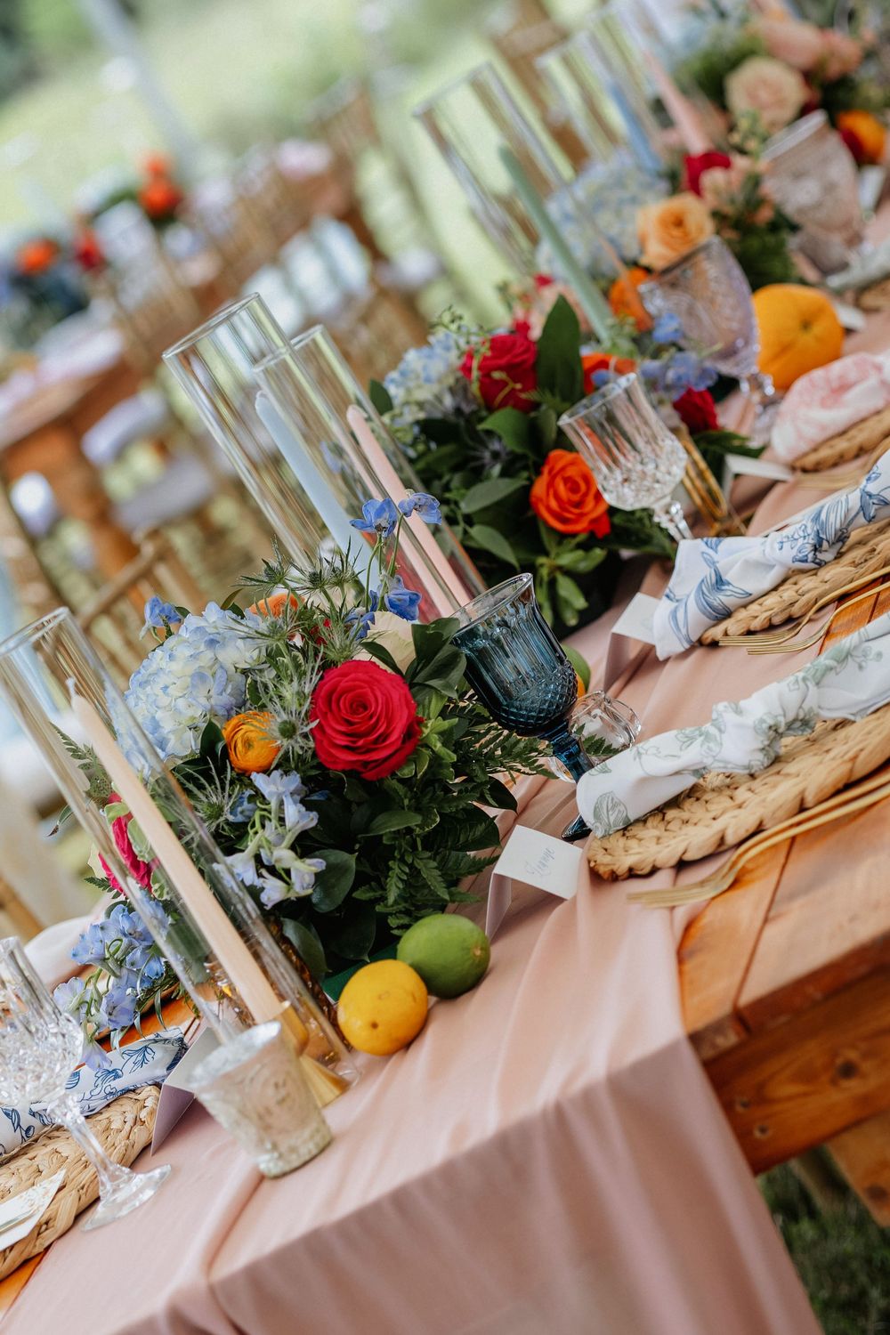 Elegant wedding reception table with colorful floral centerpieces, candles, citrus fruits, and wicker charger plates.