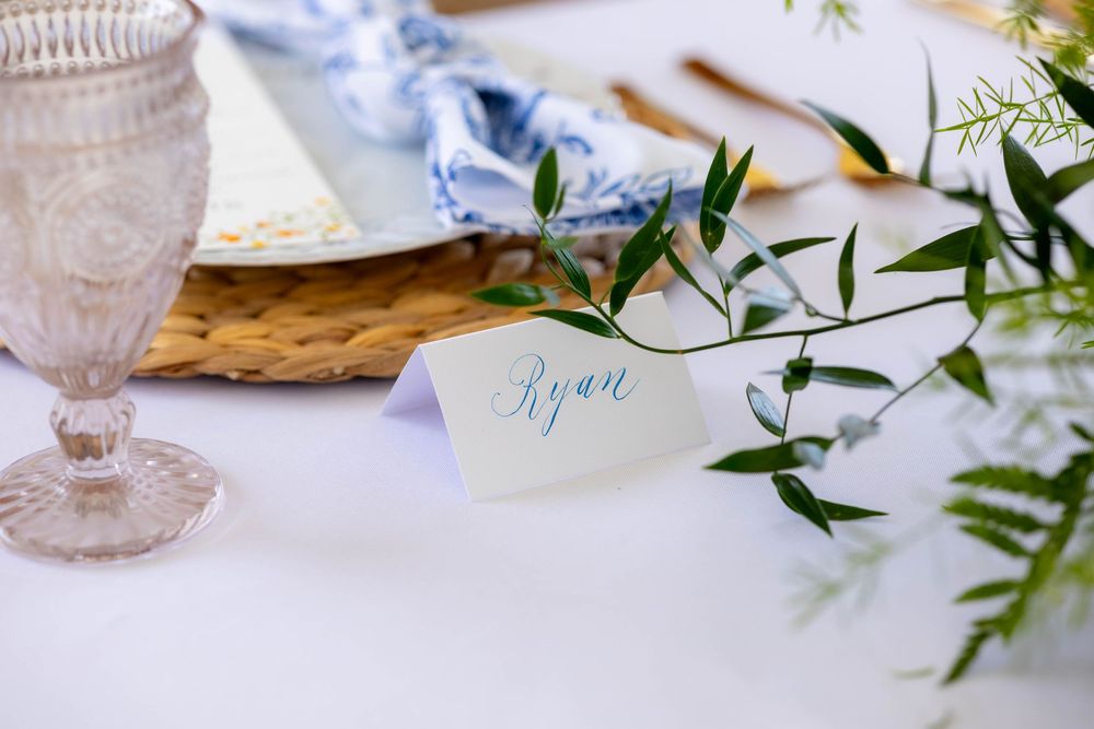 Elegant wedding place card with "Ryan" in blue calligraphy beside greenery, crystal glass, and woven charger plate.