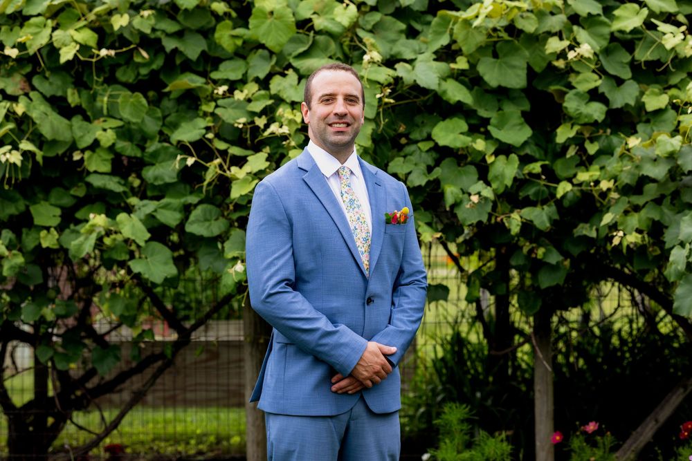 Man in blue suit with floral tie posing outdoors among lush green grapevines at a vineyard wedding.