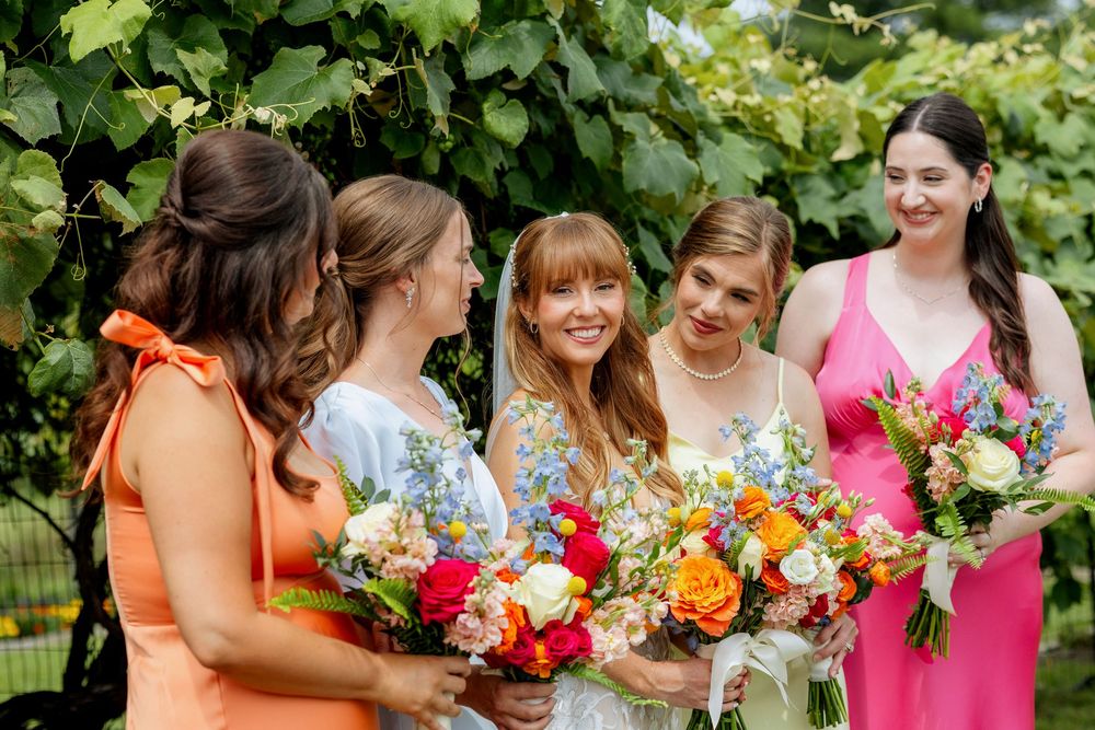 Bride and bridesmaids in colorful dresses holding vibrant floral bouquets outdoors at a vineyard wedding.
