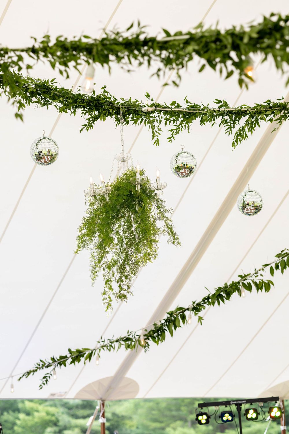 Green garland and hanging plants decorate a white tent ceiling with small glass ornaments at an outdoor event.
