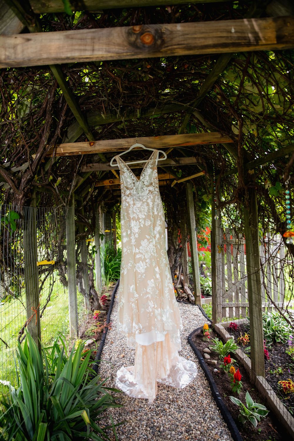 Elegant lace wedding dress hanging in a rustic garden arbor covered with lush green vines and colorful flowers.