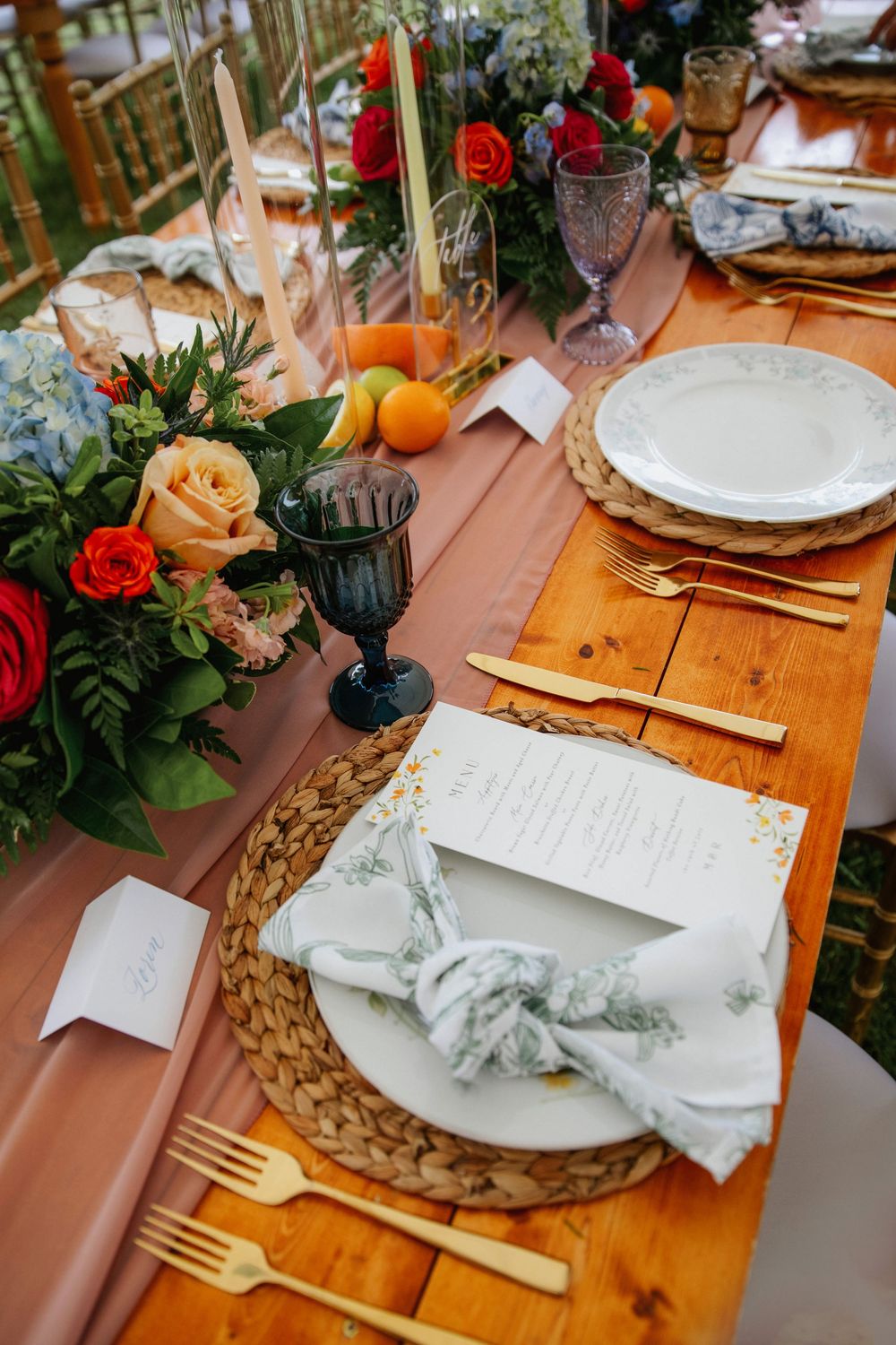 Elegant wedding table setting with gold cutlery, woven placemats, dark goblets, and colorful floral centerpiece on wooden table.