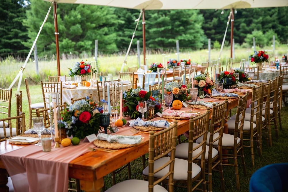 Elegant outdoor wedding reception under a tent with long wooden tables, gold chiavari chairs, and colorful floral centerpieces.