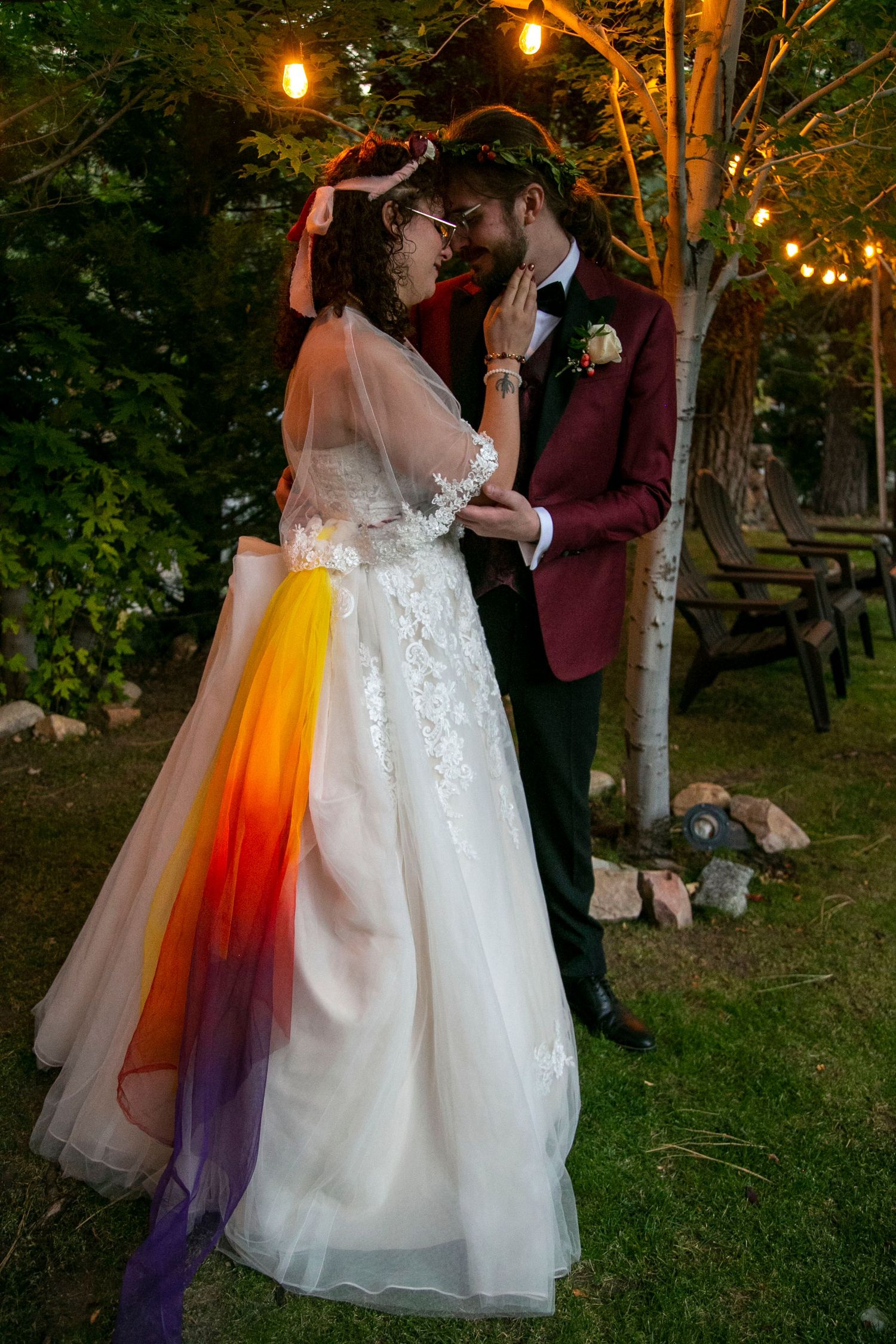 Bride in white lace gown with rainbow sash and groom in burgundy tuxedo share romantic moment outdoors at twilight.